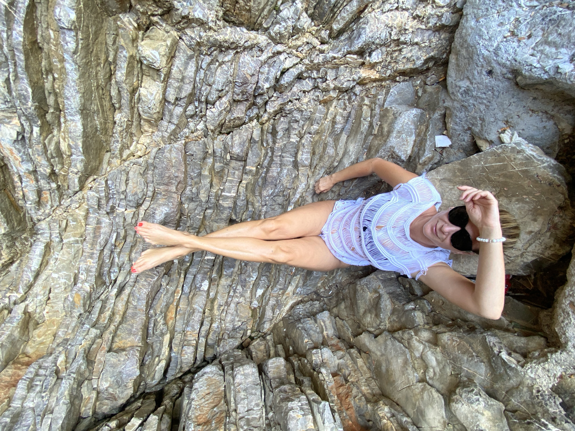 A woman is lying on her back on layered rocks, with her legs crossed and raised. She is wearing a white, sheer cover-up over a dark swimsuit and sunglasses. Her toenails are painted red. The rock formation is textured and grey, with some areas of brown and white. The location is a coastal area in Pržno, Montenegro.