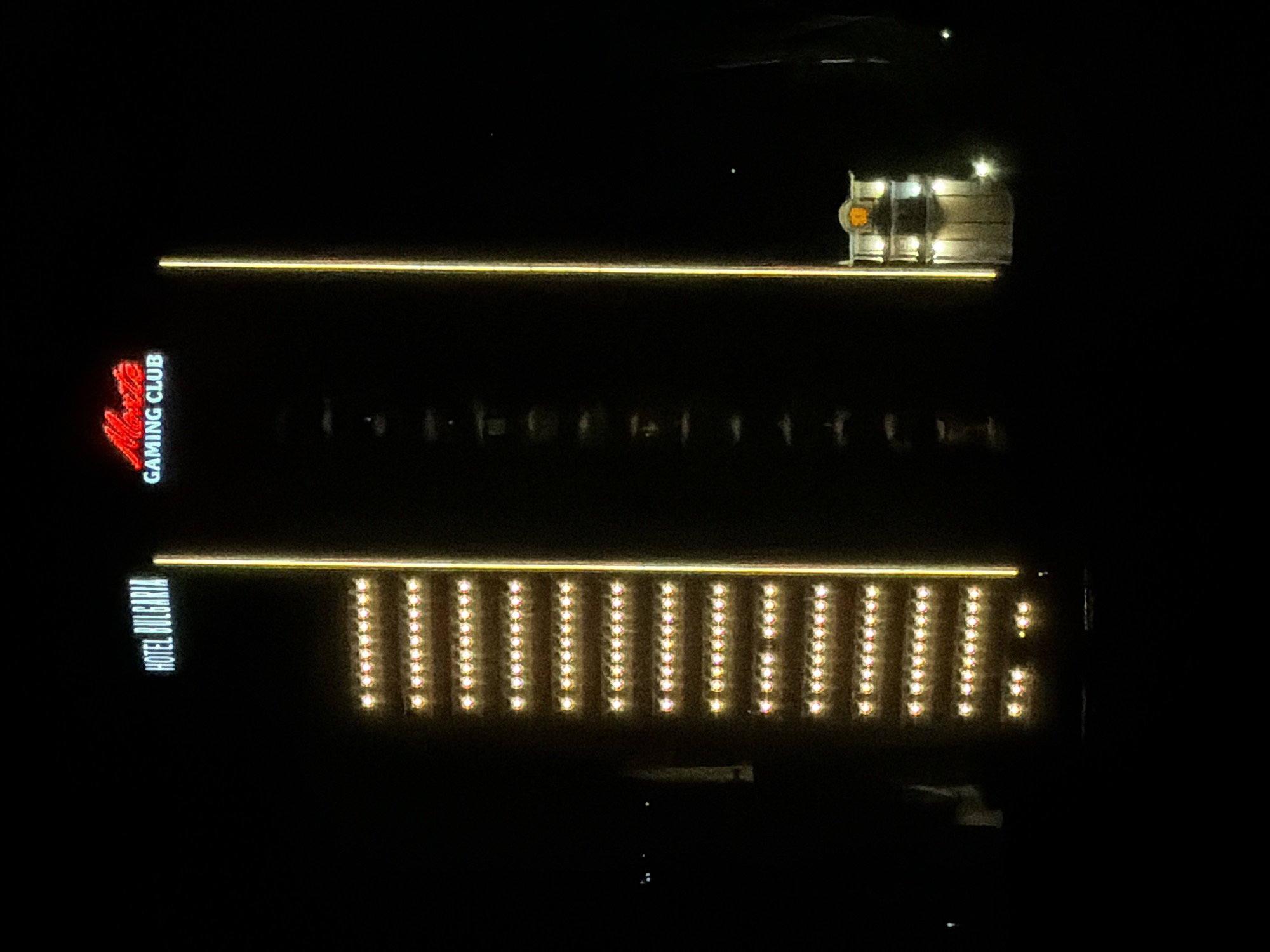 Night scene in Burgas, Bulgaria, shows a building with "HOTEL BULGARIA" signage and parallel lines of lights. Above it is another line of lights, and the signage "GAMING CLUB." A small structure with lights is at the top.