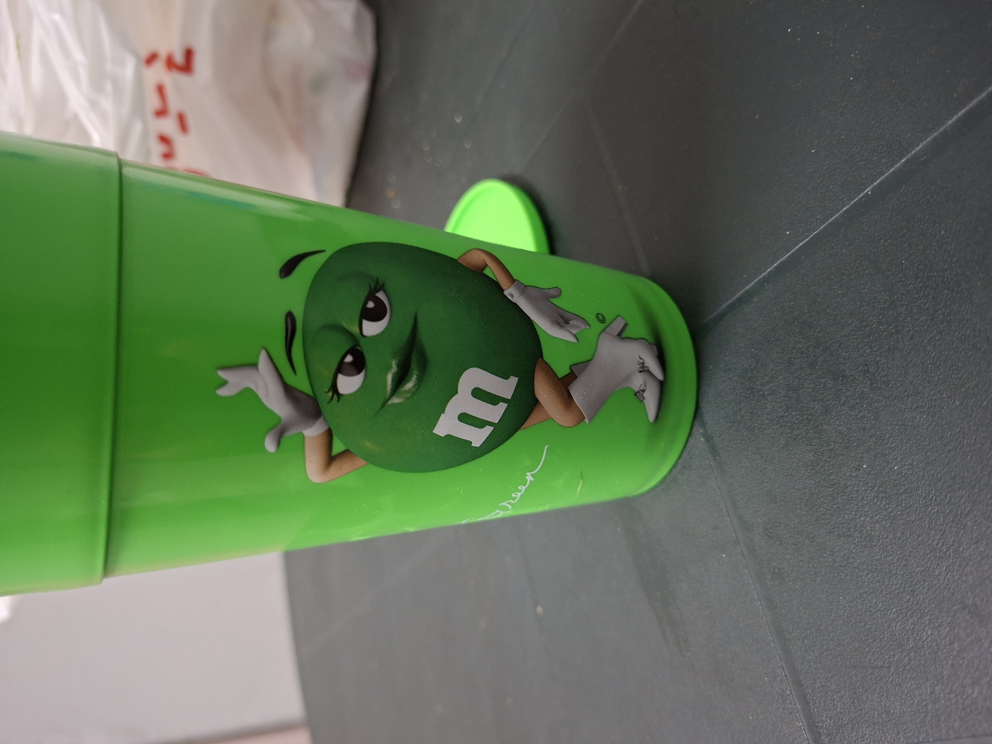 A green plastic cup with the image of the Green M&M character is on a dark gray textured surface. The cup has a green lid. In the background, a partially visible plastic bag with red writing is present. The photo does not contain sufficient data to determine a precise location within Plovdiv, Bulgaria.