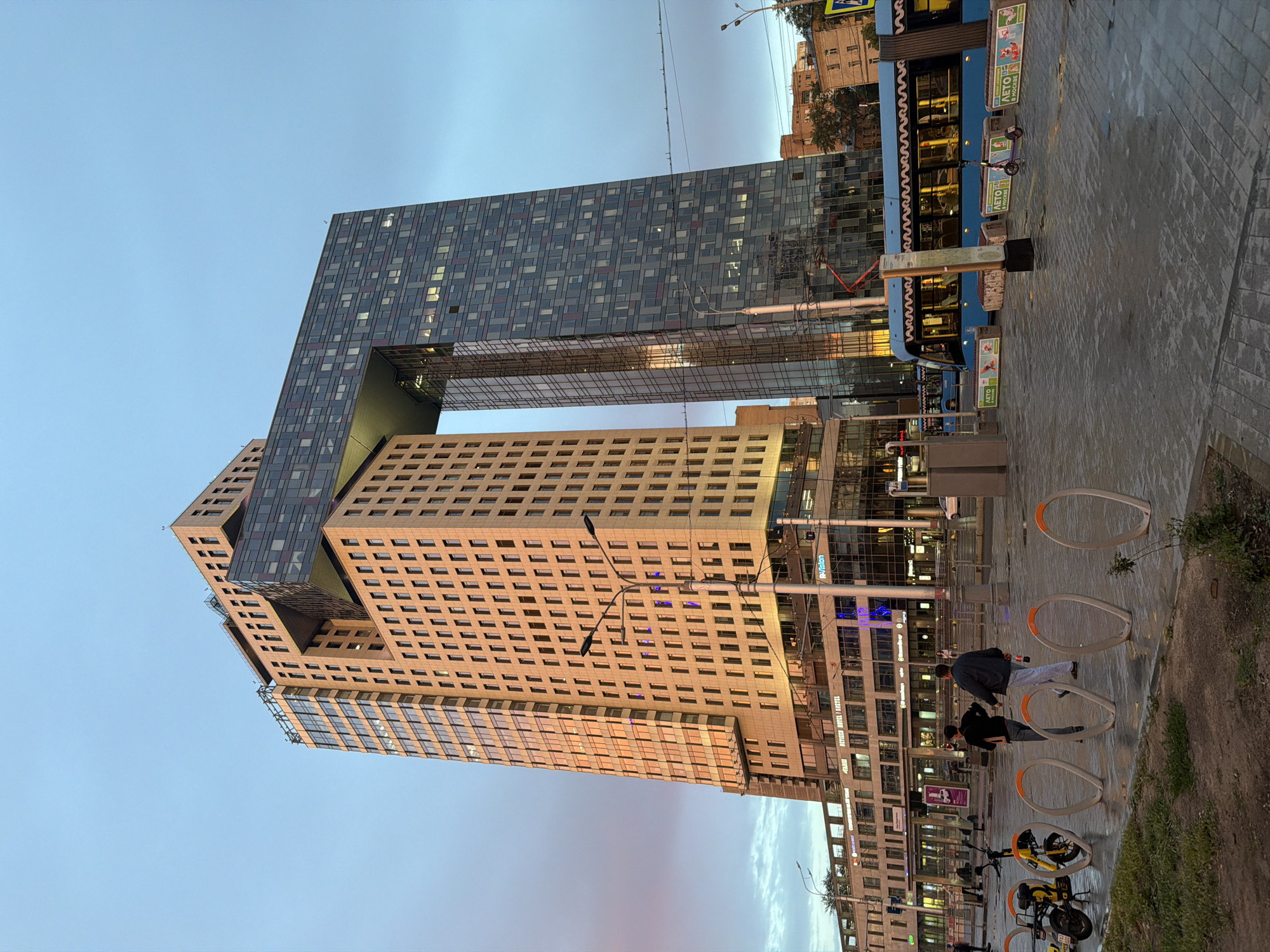 This is a vertical, outdoor photograph of a modern architectural building complex in Moscow, Russia, taken during the day. The building is uniquely designed with multiple stacked sections, one of which appears to be suspended or cantilevered over an open space. The exterior is a mix of glass facades with numerous square windows and beige, textured panels. Sunlight reflects off the building's surfaces, indicating it's likely late afternoon or early morning.
In the foreground, a paved walkway is visible, lined with bright orange bicycle racks and several bicycles, some of which are yellow. A blue tram is parked on the right side of the image, its windows reflecting the surrounding environment. Two individuals, dressed casually in dark clothing, are walking or standing near the bicycle racks.
The sky above is a clear, light blue, with a hint of pinkish hues on the horizon, suggesting the time of day. Overhead wires are strung across the top of the image. Visible text includes signs on the tram and on posters within the tram, as well as some lettering on the building facades. One sign on the tram appears to say "ЛЕТО" (Summer). The overall scene conveys an urban, contemporary atmosphere.