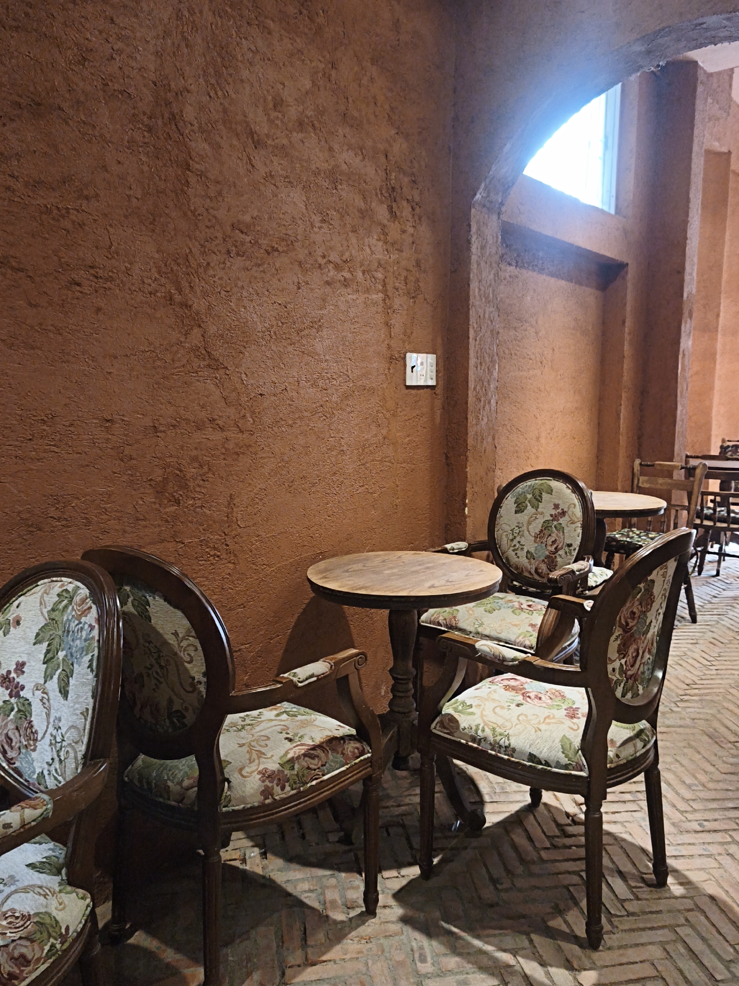 A medium shot captures an interior scene with vintage-style furniture and architectural elements. The walls are a textured, reddish-brown, and a large arched window lets in bright light from above. In the foreground, two ornate wooden chairs with floral upholstery are positioned near a small, round wooden table. The chairs have curved backs and armrests. Another similar chair is partially visible on the left. The floor is paved with reddish-brown bricks laid in a herringbone pattern. In the background, more tables and chairs, also wooden, are visible, suggesting a cafe or dining area. The overall impression is one of warmth and classic design. The location of Bac Nha Trang Ward, Vietnam is not depicted in the image.
