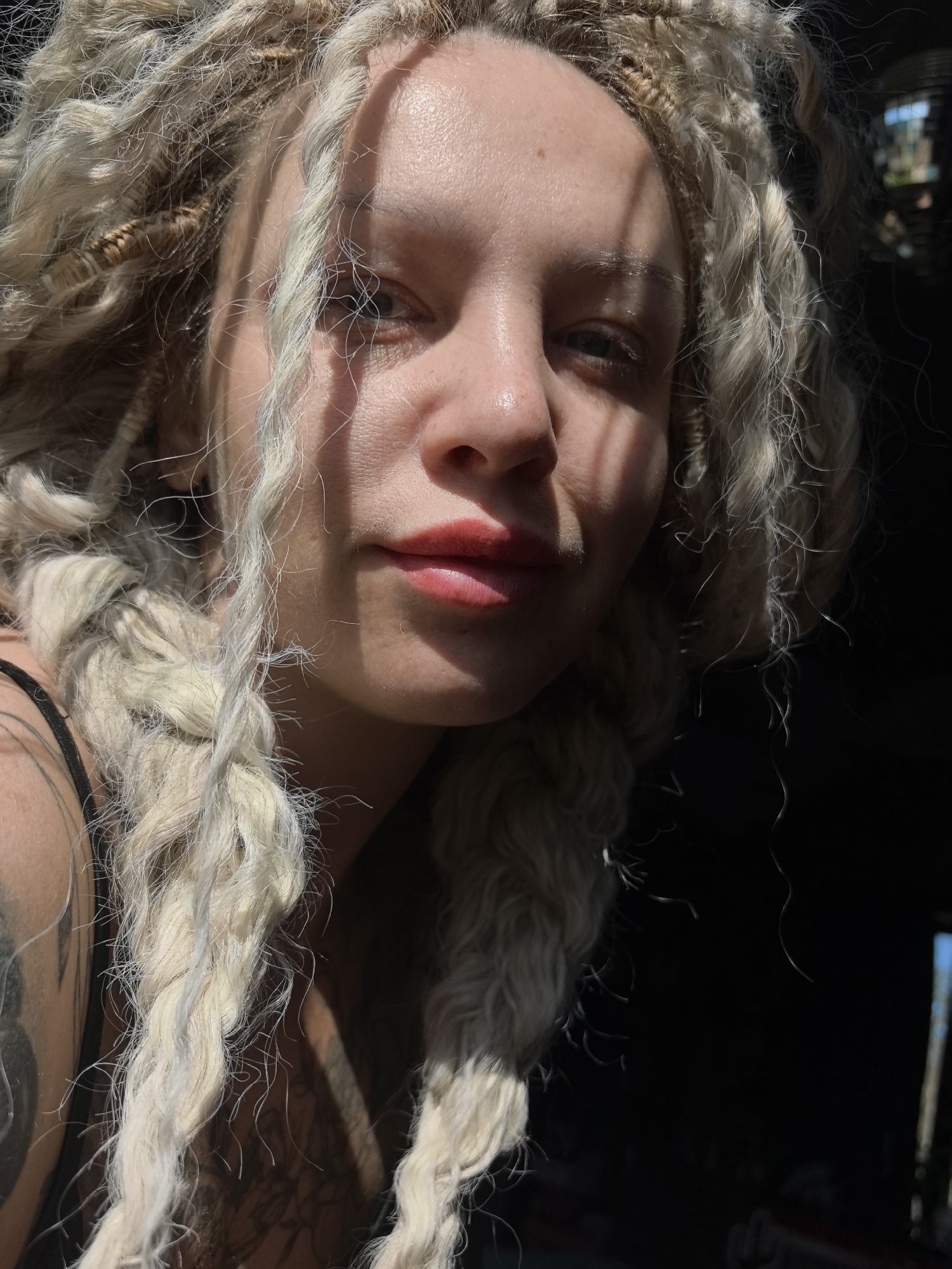 A close-up portrait features a young woman with distinctive blonde, possibly dreadlocked or heavily braided hair, which cascades around her face and shoulders. Strong, directional sunlight illuminates parts of her face, creating sharp, parallel shadows across her forehead and nose, suggesting light filtering through slatted blinds or an architectural feature. She has light-colored eyes and wears red or pink lipstick, her expression a subtle, relaxed gaze directed towards or slightly past the viewer.
Her skin appears dewy or moisturized, reflecting the bright sunlight. Visible on her left upper arm and shoulder is an intricate dark tattoo, with a hint of another tattoo on her right side. She is wearing a black strap, likely from a top, on her left shoulder.
The background is predominantly dark and out of focus, indicating an indoor setting with a powerful natural light source entering from one side. In some cropped views, a very blurry light-colored structure and a patch of blue are faintly discernible in the distant background, hinting at an exterior view beyond the dark interior space. The overall atmosphere is dramatic, characterized by the high contrast between the brightly lit subject and the deep shadows, both on her face and in the background. There is no visible text in the image. The scene suggests a sunny daytime setting.