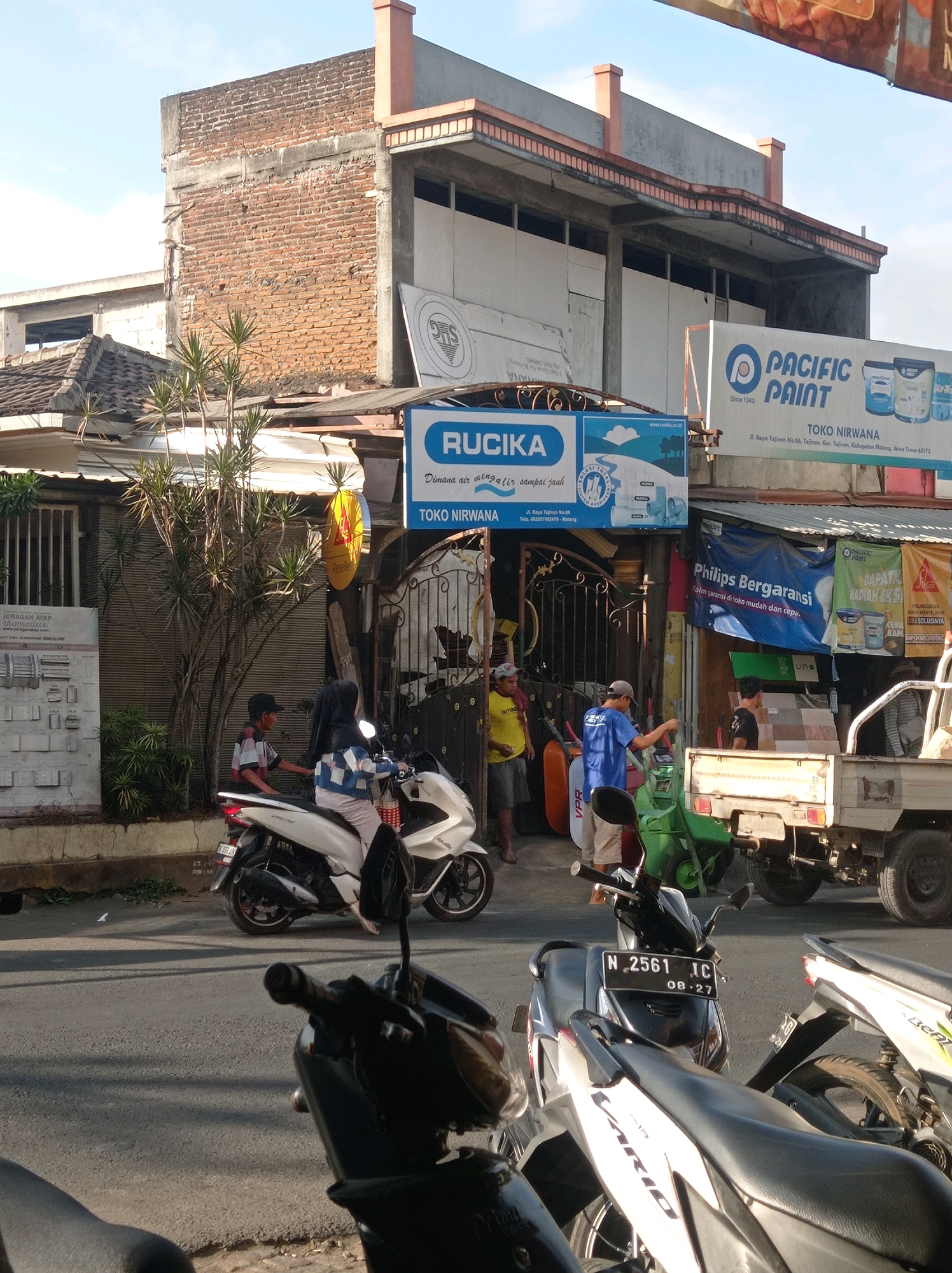 In Tajinan, Indonesia, a busy street scene unfolds under a clear sky, bathed in the light of what appears to be late afternoon. The foreground is dominated by the handlebars and seats of several parked motorcycles, their dark silhouettes contrasting with the brighter elements of the scene. A white scooter, with a rider in a patterned jacket and hijab, is parked in the middle ground, angled towards the left. To its right, a young boy wearing a striped shirt and a cap stands near a green scooter.
Behind the vehicles, a row of shops lines the street. A prominent blue sign for "RUCiKA" is visible, advertising plumbing solutions, and below it, a sign for "TOKO NIRWANA". Adjacent to this is a larger sign for "PACIFIC PAINT". Several people are seen interacting in front of these shops. One man in a blue shirt and cap stands near a white truck, while another man in a yellow shirt and shorts stands to his left. A truck with a light-colored bed is parked in front of the shops, its rear tire on the right side of the frame. The buildings themselves show a mix of architectural styles, with brickwork and concrete elements visible, suggesting a blend of older and newer construction. The street surface is asphalt, and patches of dry grass are visible along the roadside. The overall impression is one of everyday commerce and activity in a semi-urban Indonesian setting.