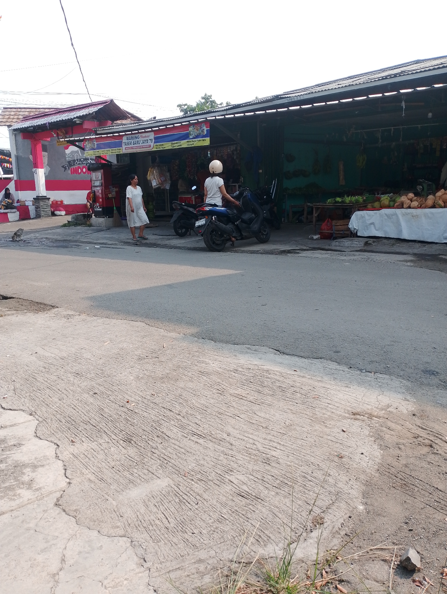 A wide shot captures a roadside market in Bogor, Indonesia, under a clear sky. In the foreground, a cracked and weathered concrete road dominates the lower half of the image. To the left, a person on a motorcycle, wearing a white helmet and a white t-shirt, is stopped. Adjacent to this, a woman in a white dress walks away from the camera. Behind her, a row of market stalls features a selection of fruits, including coconuts and bananas, displayed on tables covered with white cloths. The stalls are covered by a corrugated metal roof supported by wooden poles. A banner above the stalls reads "WARUNG MAKAN TASER BARU JAYA 76". To the far left, a small building with red and white striped walls has an image of a man's face painted on it. The scene appears to be a typical street market in a populated area.