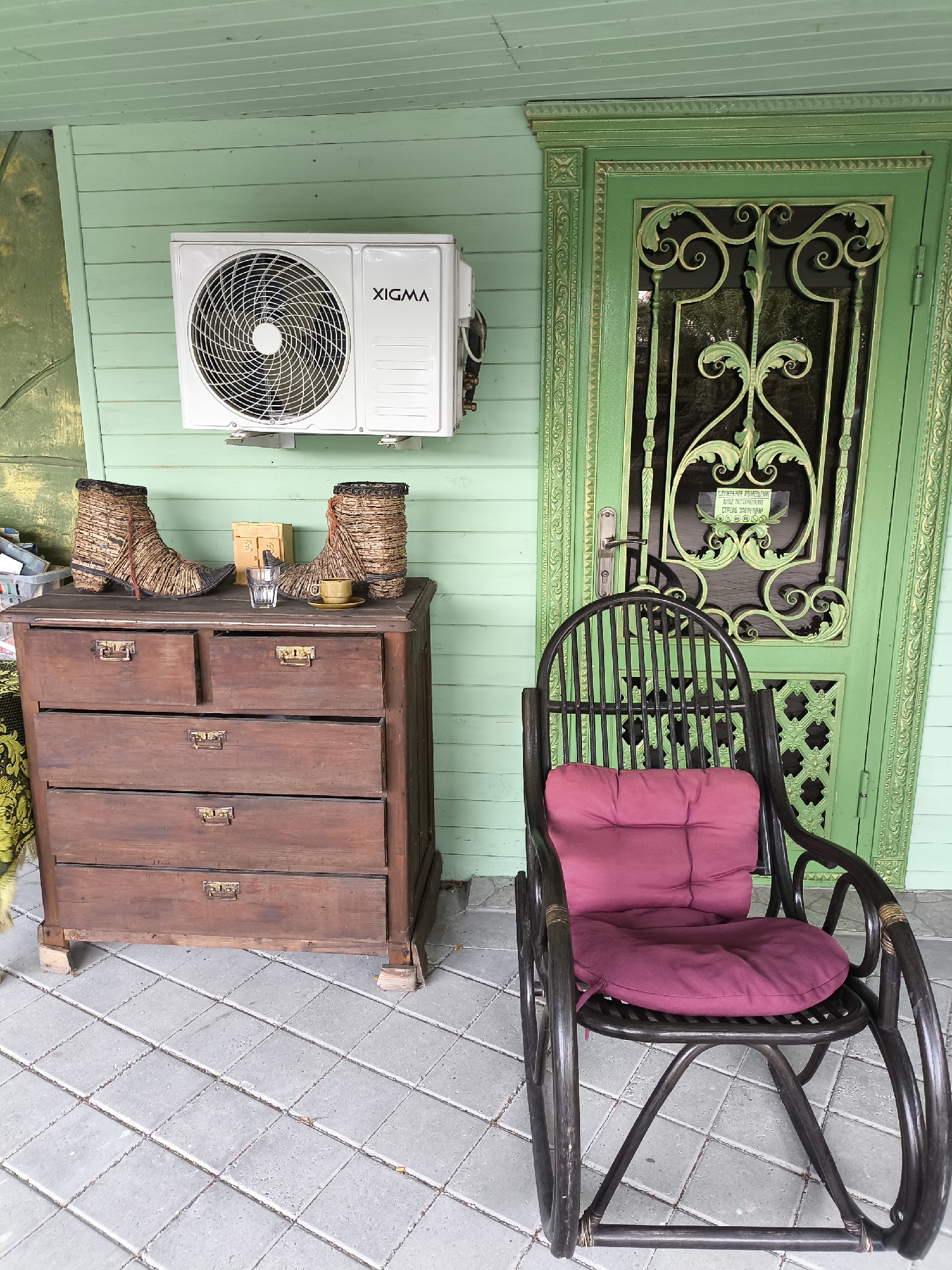 In Borgustanskaya, Russia, a tranquil scene unfolds on a covered porch. A wooden dresser, stained a rich mahogany, stands against a pale green wooden wall adorned with a white XIGMA air conditioning unit. On the dresser, two woven, boot-like decorative items sit on either side of a small glass of water and a miniature teacup with a saucer, all illuminated by natural light. To the right, a dark wicker rocking chair, cushioned with a vibrant pink pad, faces a weathered green door with intricate metalwork. The floor is tiled with light gray squares.