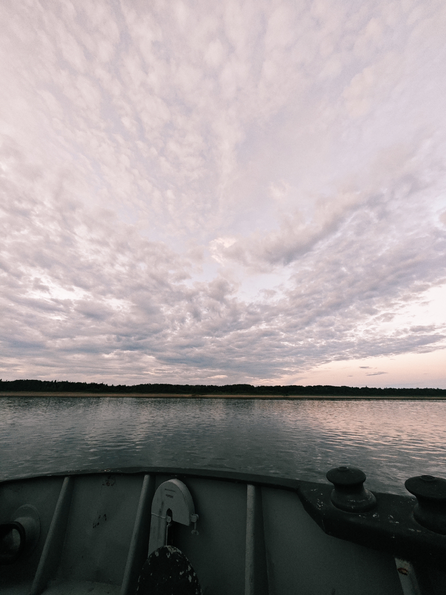 The image shows a view from the deck of a boat traveling on a wide body of water. The foreground is dominated by the gray metal deck of the boat, with visible bollards and some equipment. The water is calm, reflecting the muted colors of the sky. A dark, tree-lined shore is visible in the middle ground, stretching horizontally across the frame. Above the horizon, the sky is filled with a vast expanse of clouds, tinged with soft pink and gray hues, suggesting either sunrise or sunset. The overall mood is peaceful and serene, evoking a sense of quiet travel. The text "ТОРМОЗИ" is visible on a white label attached to a piece of equipment on the boat deck. Based on the location context of Nikulino, Russia, the scene likely depicts a calm journey on a river or lake in the early morning or late evening.