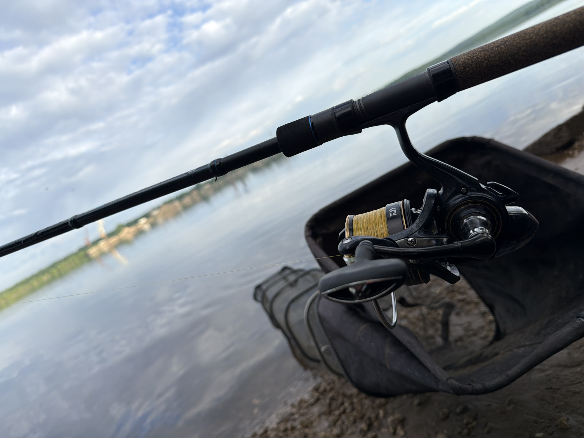 The image shows a fishing rod and reel positioned over a landing net on the bank of a body of water. The reel is spooled with yellow line. In the background, a blurred cityscape can be seen across the water. The scene appears to be in Chaikovsky, Russia, based on the architectural style and landscape visible in the background. The sky is overcast with light clouds. The immediate foreground consists of what appears to be muddy shoreline.