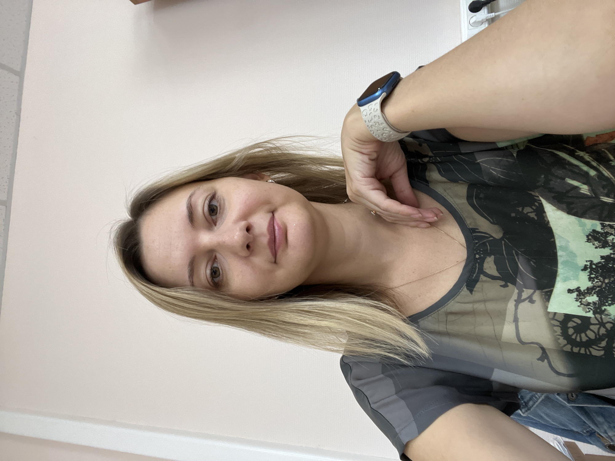 A medium shot, taken from a slightly high angle, shows a woman with blonde hair and brown eyes. She is wearing a grey t-shirt with a nature-themed print and a necklace. On her left wrist, she wears a smartwatch with a white band patterned with leopard spots. Her left arm is bent, with her hand resting on her chest. She is indoors, with a pale pink wall and a white ceiling visible behind her. To her left, a portion of a white wall and a doorway are visible. To her right, a power strip with two plugs is mounted on the wall. The scene appears to be in an office or home environment.