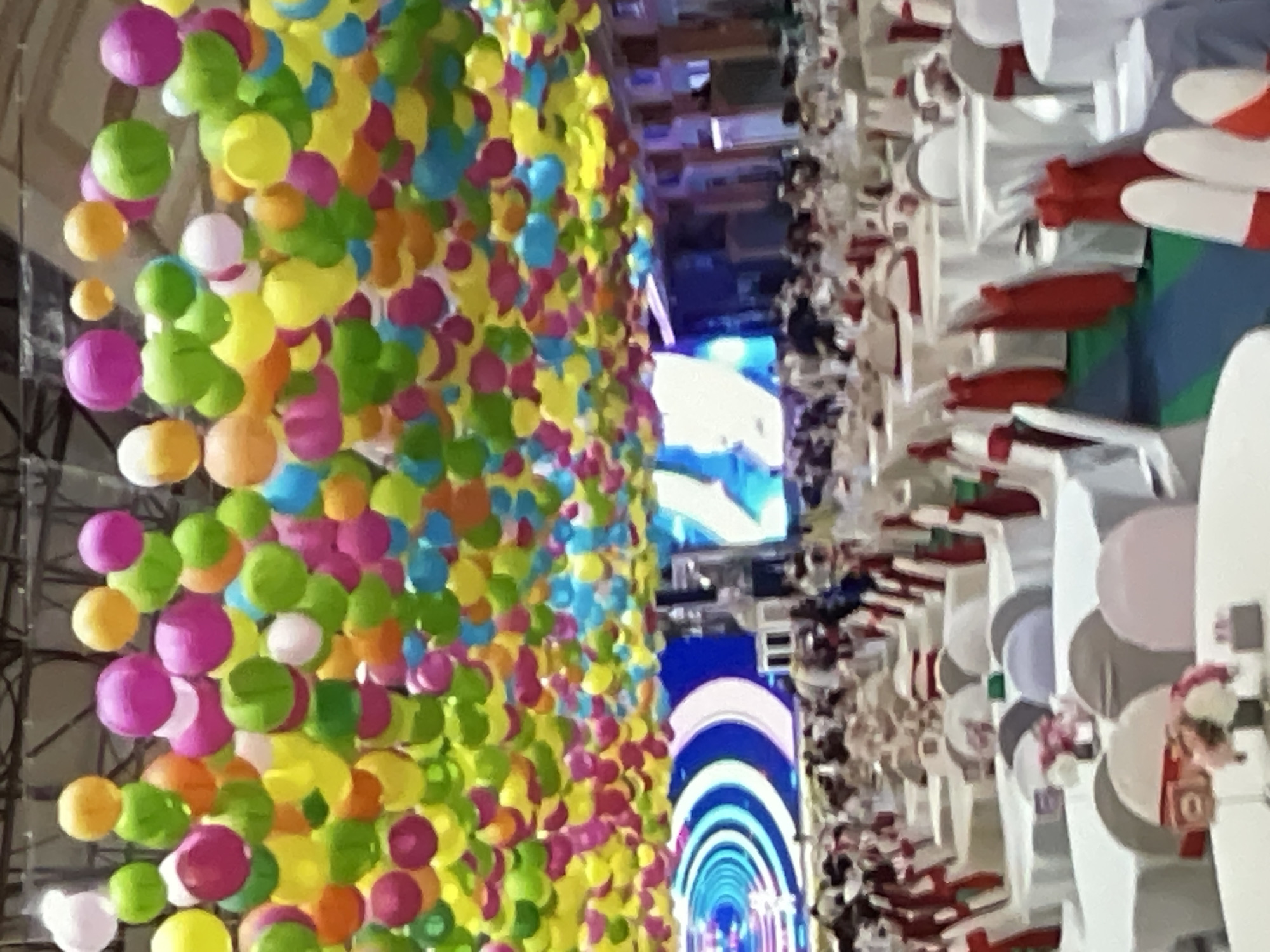 The image depicts an indoor event space, likely a banquet hall or convention center, with a vibrant ceiling adorned with numerous colorful, round lanterns in shades of green, yellow, pink, orange, and blue. These lanterns are densely clustered and create a festive canopy above the scene.
Below the lanterns, the space is set up for an event with many round tables covered in white tablecloths. The chairs around the tables are mostly white, many with red sashes tied around them, adding a touch of color to the setting. Small floral arrangements and other decorative items are visible on some of the tables.
In the background, a large screen or display is visible, showing a blue and white circular graphic, possibly a logo or part of a presentation. There are also figures of people, appearing as a crowd, moving or standing in the background, though they are not clearly discernible due to the distance and the image's resolution. The overall impression is of a well-decorated event venue, possibly a celebration, conference, or performance. The location, Pattaya City, Thailand, suggests a tropical or subtropical setting, which might influence the choice of vibrant colors and a festive atmosphere for an event.