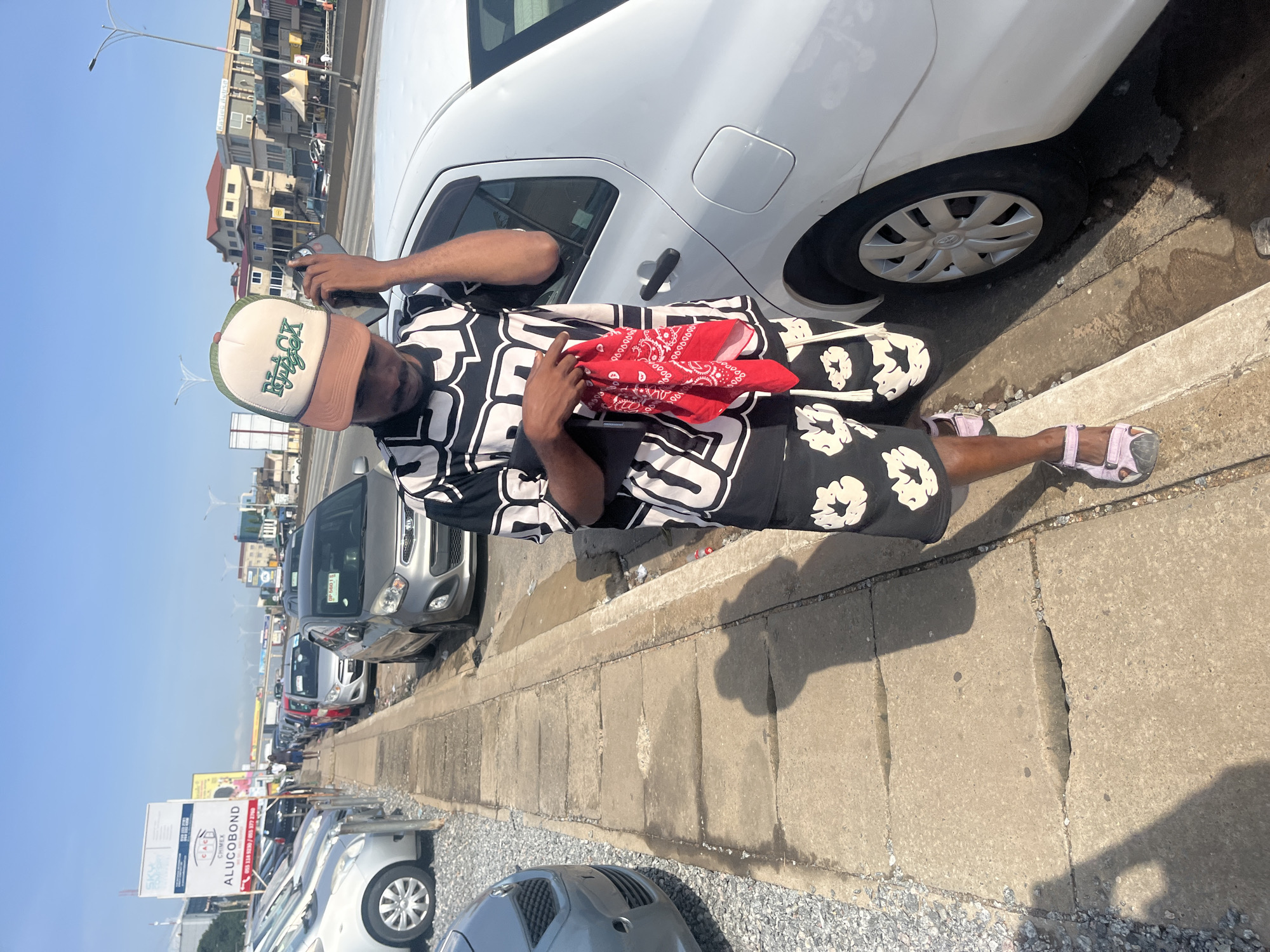 A Black male is standing on a sidewalk next to a white car. He is wearing a black and white patterned t-shirt, black shorts with white patterns, a green and brown trucker hat, and pink sandals. He is holding a red bandana in his left hand and talking on a cell phone held to his right ear. The sidewalk is made of concrete and is cracked in several places. A row of cars is parked on the left side of the sidewalk. In the background, there is a highway with traffic, buildings, and a blue sky. The scene appears to be in Accra, Ghana, indicated by the presence of an "ALUCOBOND" advertisement which is common in Ghana, and the architectural style of the buildings.