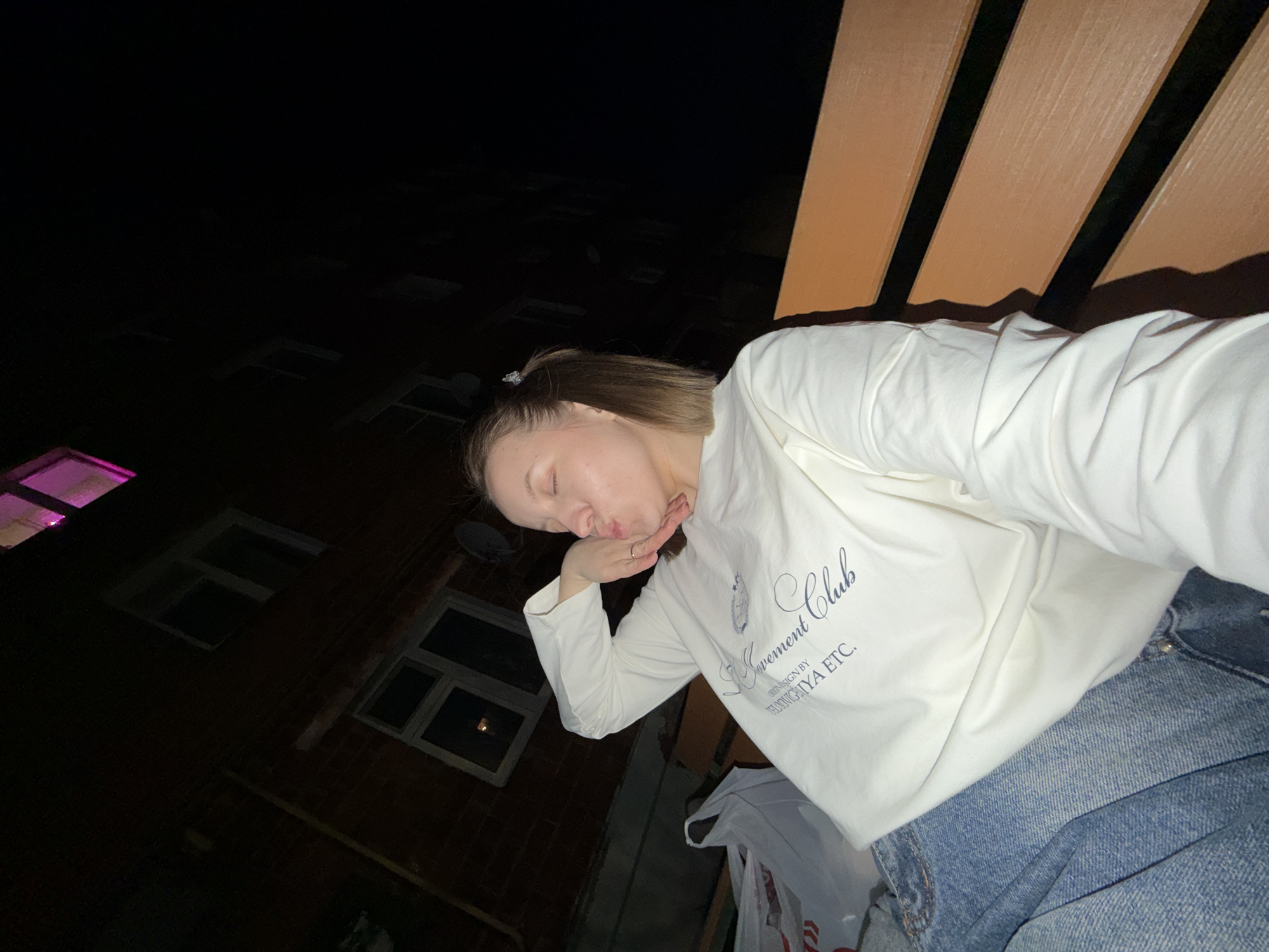A woman is seated, leaning against a wooden-slatted structure, possibly a bench or fence. She's wearing a white long-sleeved shirt and jeans, and appears to be blowing a kiss towards the camera. Behind her, there's a building with visible windows, one of which has a pink light. The scene seems to be at night, with limited ambient light. The image does not specify the city of Reshetnikovo, Russia.