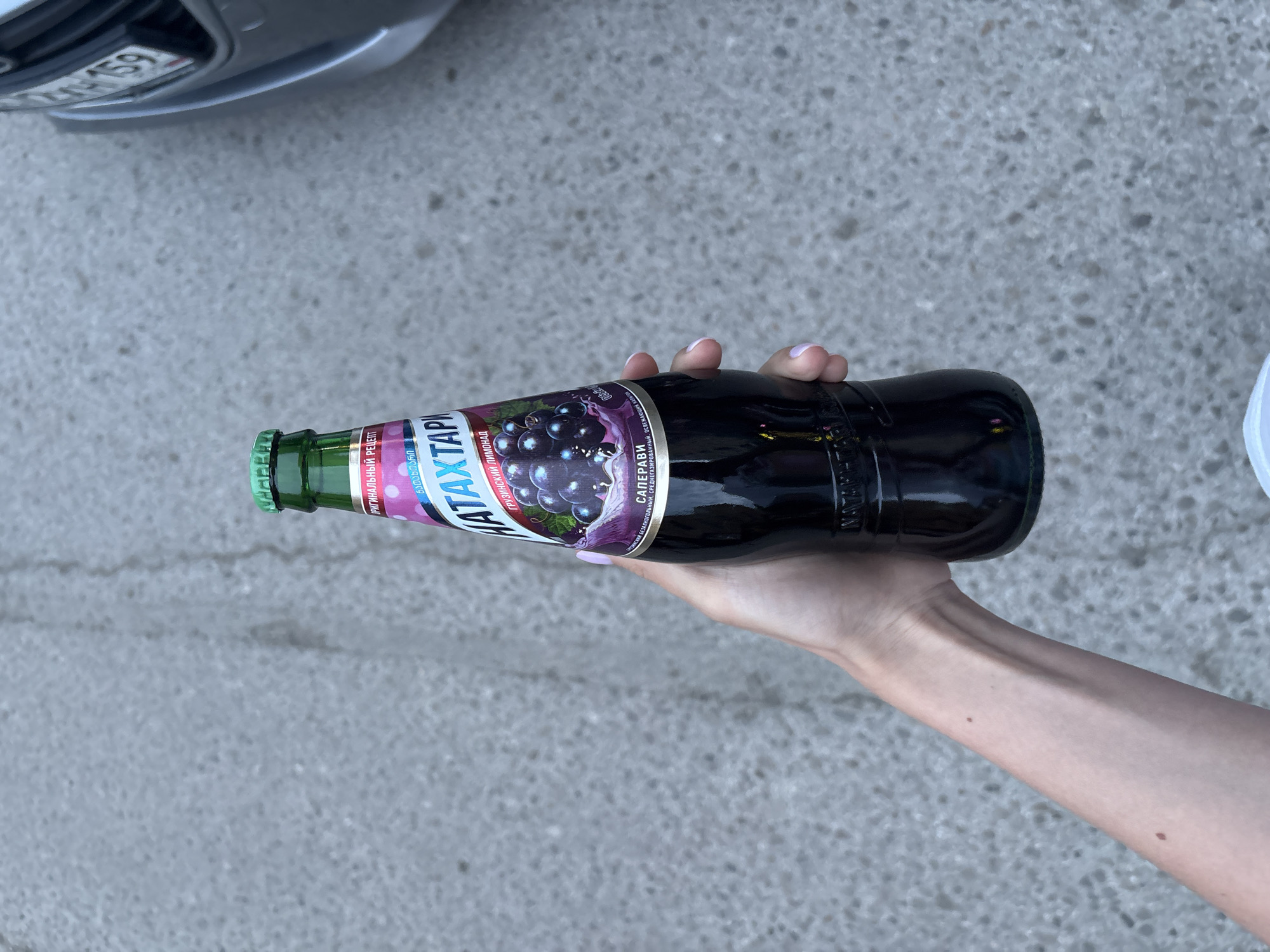 A hand is holding a bottle of Natakhtari Saperavi grape soda. The bottle is dark and has a green cap. The label on the bottle is in Russian and Georgian. The background is a paved surface, potentially a sidewalk or street. Part of a car bumper with the license plate “KH 159” is visible in the upper left corner. A small portion of a white garment is visible on the right edge. The scene likely takes place in Russia, given the Russian license plate and the writing on the label. It could be in Perm, Russia, as the bottle is in Perm, based on the location provided.