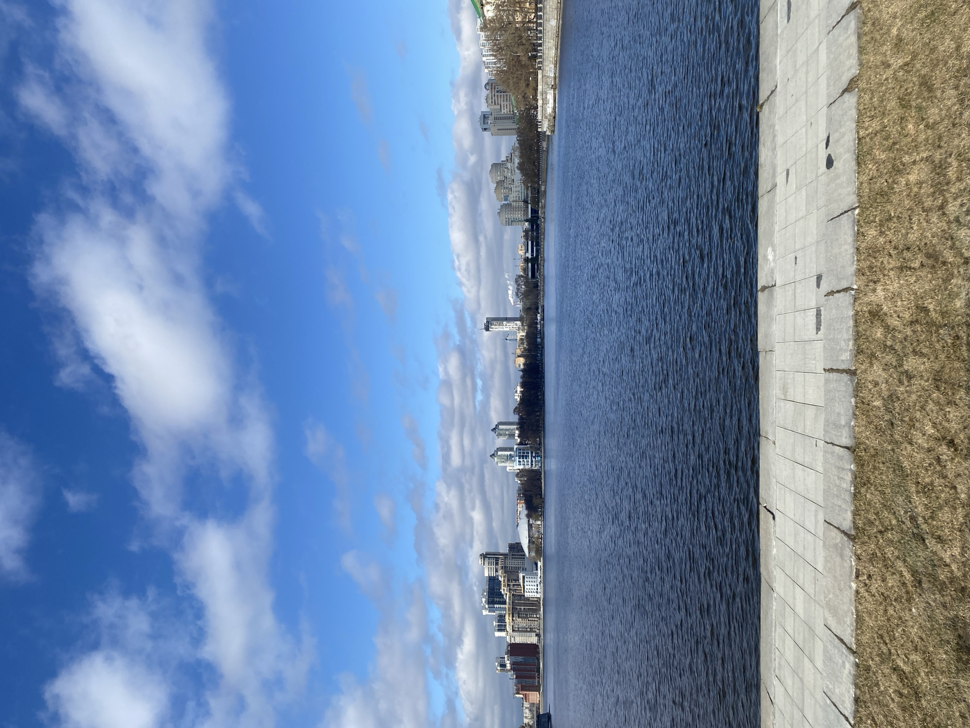 The image shows a view along the Iset River in Yekaterinburg, Russia. The scene is divided into three distinct areas. On the left is a blue sky with scattered white clouds. In the middle is the dark blue Iset River. On the right is a gray concrete embankment with dry grass adjacent to it. Buildings are visible along the riverbank.