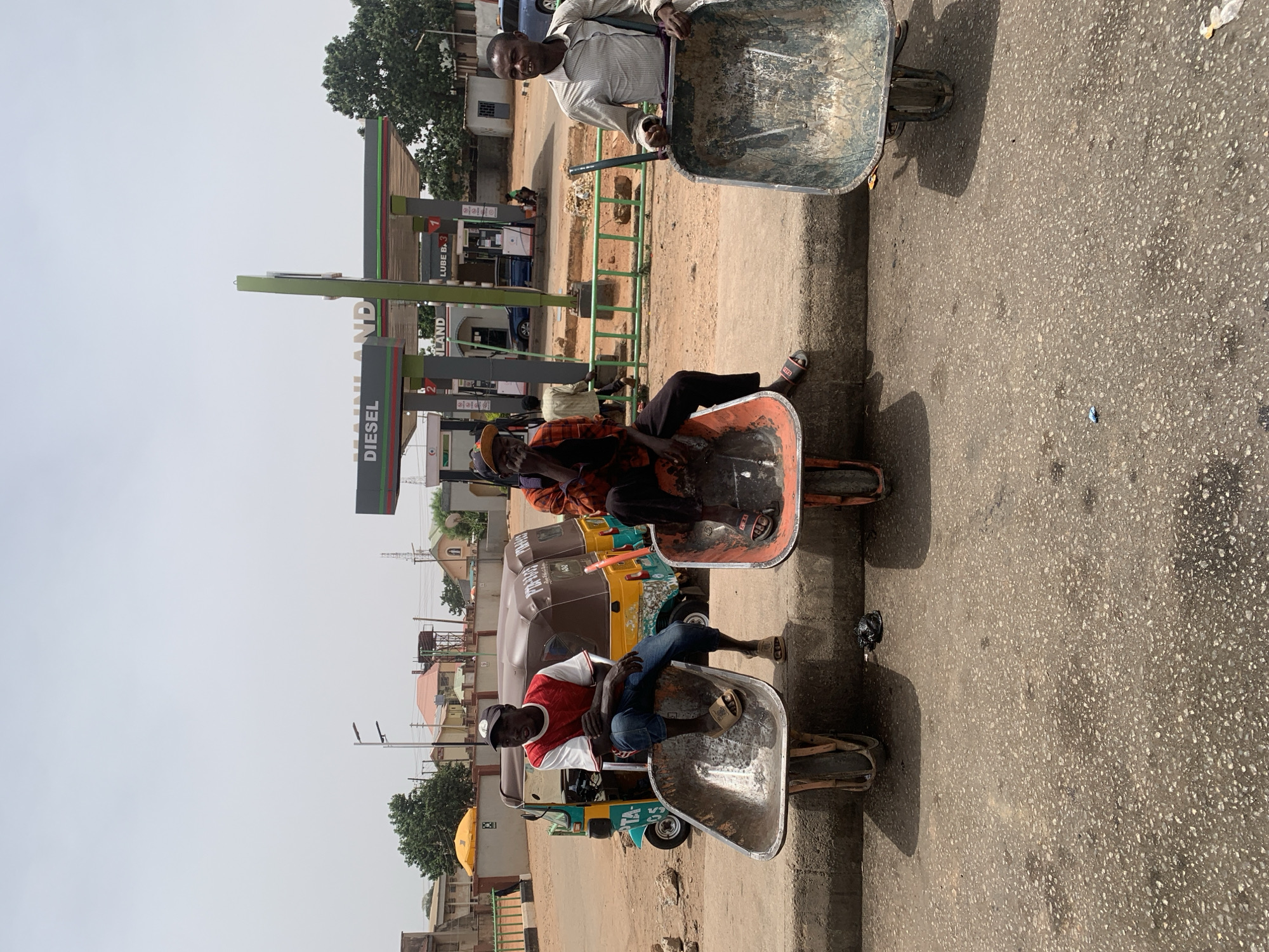 The image shows three men sitting in wheelbarrows, lined up on a paved surface, likely a road. Behind the men, there are several auto-rickshaws parked. In the background, there is a gas station branded "MAINLAND" with "DIESEL" visible. The environment suggests an urban setting in Jos, Nigeria, based on the vehicles, gas station, and architectural styles in the background. The men are interacting by being positioned next to each other, and their actions involve sitting in wheelbarrows, possibly as a form of entertainment or transportation. The surrounding environment includes buildings, trees, and road infrastructure, indicating a populated urban area.