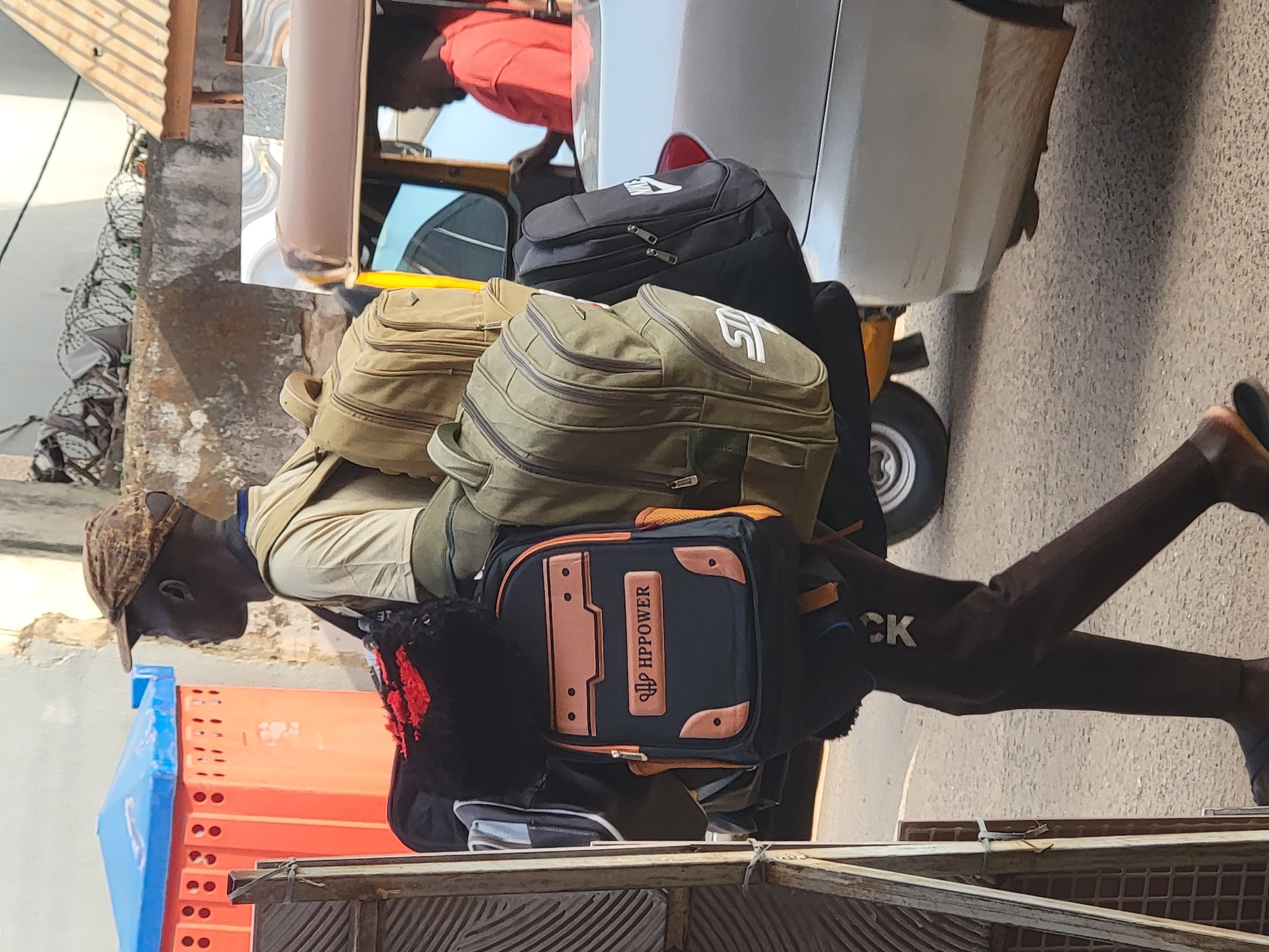 A man with dark skin is carrying a large stack of backpacks on a cart in Maiburiji, Nigeria. He wears a tan shirt, brown pants, and a patterned baseball cap. The cart is situated on a paved street, next to a building with orange and blue elements. A white vehicle, possibly a taxi or similar service, is parked nearby with another person inside. Barbed wire is visible atop a wall in the background.