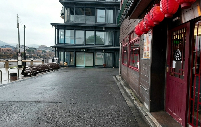 The image displays an urban waterside scene in Arashiyama, Kyoto, Japan, under overcast conditions. The foreground consists of wet, dark asphalt pavement. To the right, a building with dark reddish-brown horizontal siding features multiple red-framed windows and a dark red entrance door with a white, stylized plant emblem. Seven red paper lanterns with black Japanese characters hang in a row from an awning above this entrance. A poster display is visible beside the door.
To the left of the pavement, an embankment borders a visible body of water (likely a river). Along this embankment, a long, dark, textured metal bench, appearing as two curved sections, is positioned. A tall utility pole, an information sign, and a metal waste receptacle are also present.
In the midground, a modern, multi-story building with a dark grey facade and numerous glass windows stands. The name "ゆらり" (Yurari) is displayed on its ground floor. The background reveals distant buildings across the waterway, set against a backdrop of rolling hills under a cloudy sky. No individuals or active subjects are visible.