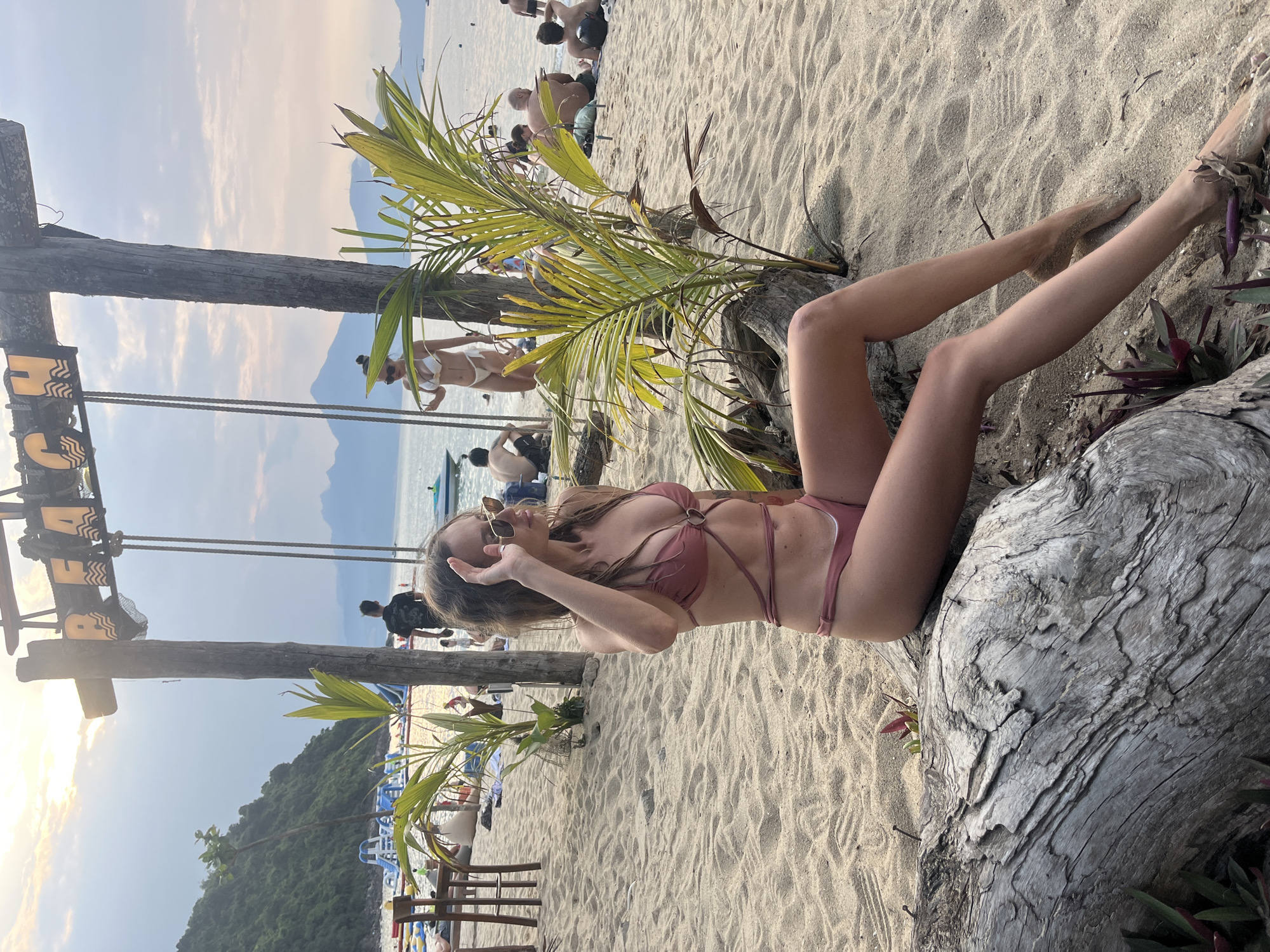 A young woman in a rust-colored bikini sits on a weathered tree trunk on a sandy beach in Đà Nẵng, Vietnam. The background includes trees, other beachgoers, and a view of the water. The time of day appears to be late afternoon or early evening, given the position of the sun.