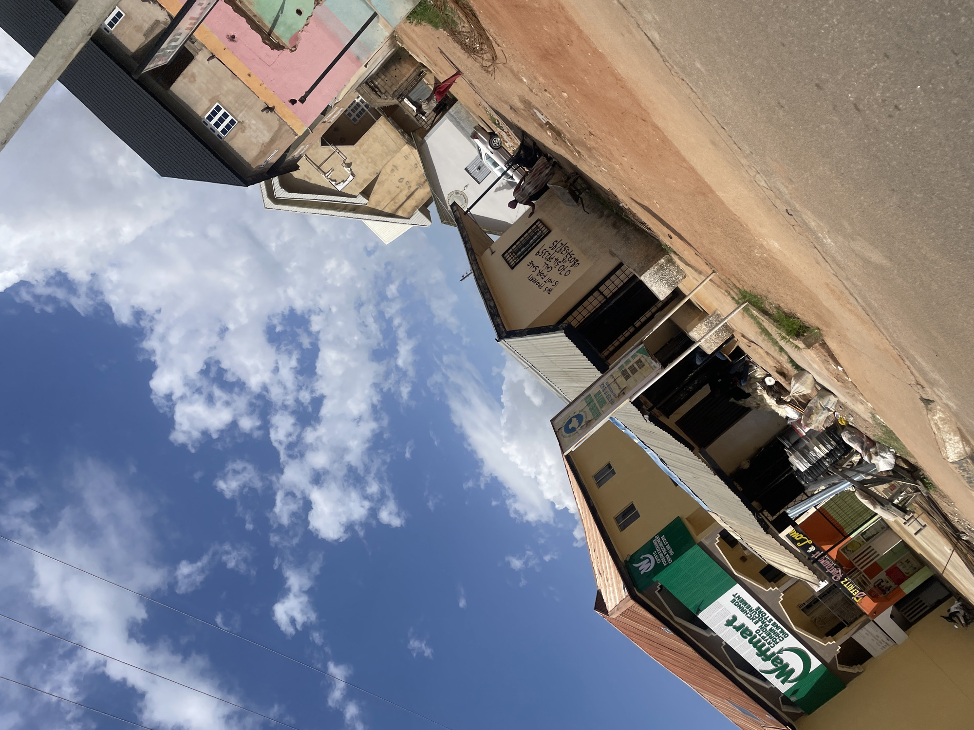 The image captures a street scene in Bar Jirgi Summa, Nigeria, under a partly cloudy blue sky. The buildings are predominantly two to three stories high, with varied architectural styles and paint colors, including shades of beige, yellow, pink, and green. Some buildings appear to be under construction or in disrepair.
On the left side of the street, a yellow building features signs for "Waffmart," advertising Crypto Foreign Exchange, China Procurement, and an Online Store. Another green sign above it also displays the "Waffmart" logo and text. Adjacent to this, a smaller storefront with an orange and green facade is visible, with the name "DeFritz Outrageous & Lou..." partially legible.
In front of the buildings, there's a dirt and gravel pathway, with some grass growing along the edges. Piles of trash and building materials are scattered on the ground, indicating ongoing activity or neglect. A silver car is parked on the street near a white building. Two individuals are present in the mid-ground; one is wearing a red garment and appears to be walking, while another, dressed in a patterned robe, is standing near a wall with graffiti that reads "THIS PROPERTY IS NOT FOR SALE CALL...". A motorcycle is parked on the lower left of the image.
Several power lines are visible traversing the sky, and a concrete utility pole stands on the right side of the frame. The overall impression is of a commercial area with a mix of established businesses and developing infrastructure. The image is taken at an angle, suggesting the photographer was looking upwards and sideways.