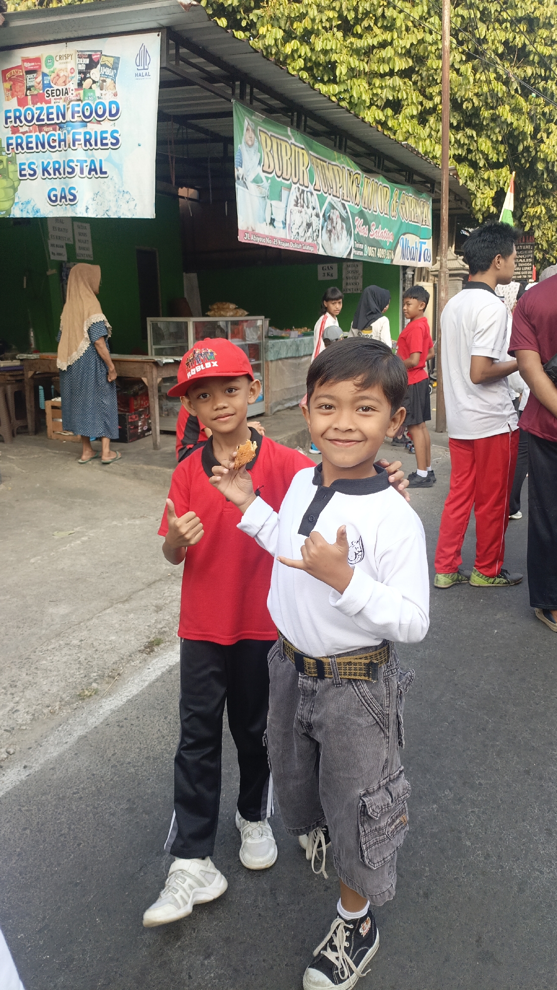 The image depicts an outdoor scene in front of a food stall in Salatiga, Indonesia. Two young boys are prominently featured in the foreground, smiling at the camera. The boy on the left wears a red cap and a red t-shirt, holding a piece of food and giving a thumbs-up. The boy on the right wears a white long-sleeved shirt and grey cargo shorts, making a hand gesture. In the background, several other individuals are visible, along with banners displaying food items and prices. One banner, with text in Indonesian, advertises "BUBUR AMPLANG KOTOR & GORENGAN" and mentions the location as "Jl. Abyoso No. 25 Krajan Dukuh Salatiga". Another banner on the left lists "FROZEN FOOD FRENCH FRIES ES KRISTAL GAS". The environment appears to be a street with a paved surface and a building with a canopy providing shade. The overall atmosphere is casual and lively, suggesting a marketplace or street food area.
