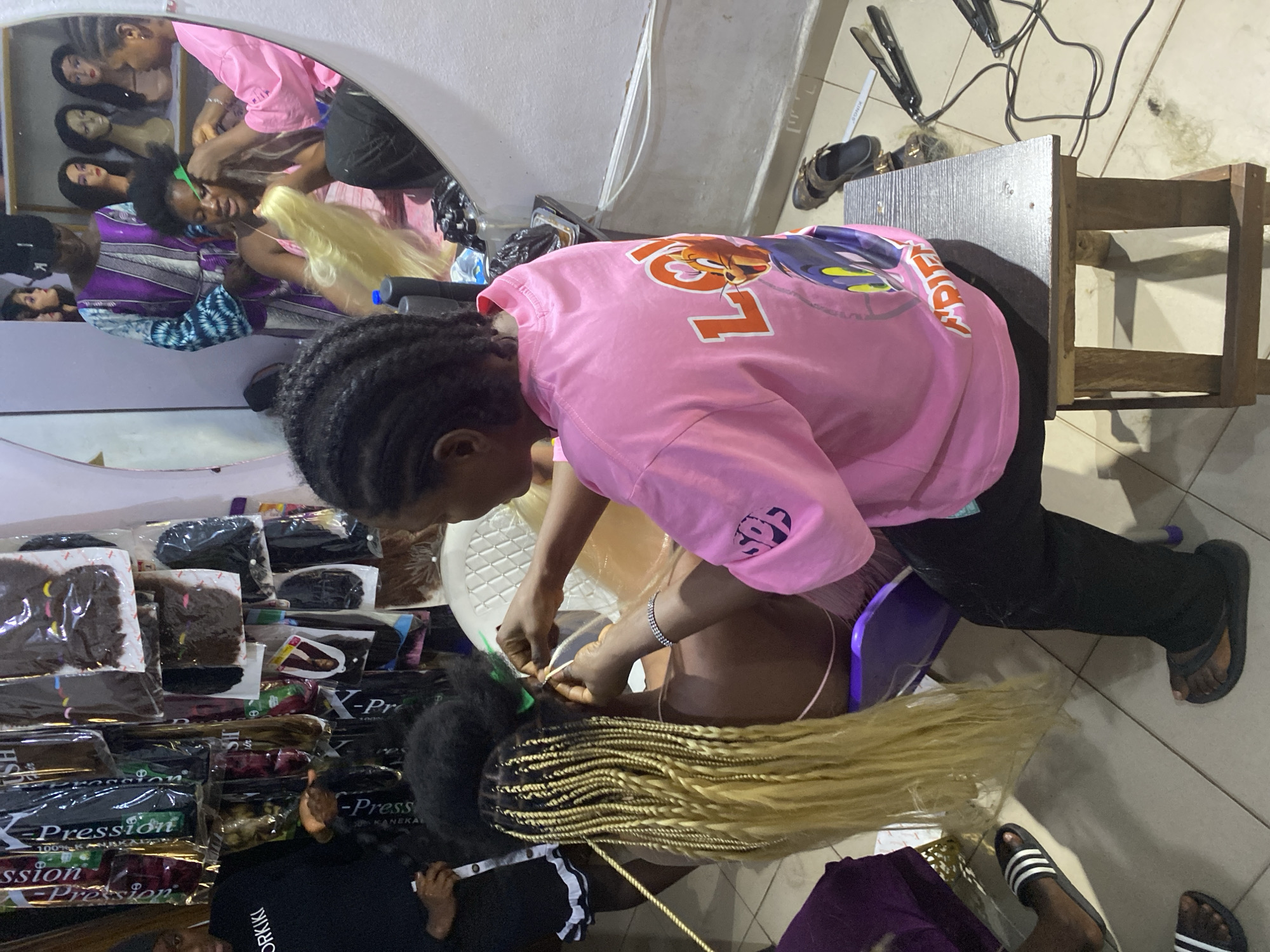 In a brightly lit establishment in Bukuru, Nigeria, a woman wearing a pink graphic t-shirt and black pants is braiding another woman's hair. The customer is seated on a small purple stool and has long, blonde braids already completed. The stylist is working on the customer's natural black hair, adding more blonde braiding hair.
Behind the stylist is a wall lined with various packages of hair extensions. In the background, a mirror reflects the scene, showing additional mannequins with wigs. On the floor, near a wooden stool, are a hair straightener, shoes, and electrical cords. The floor tiles are light colored.