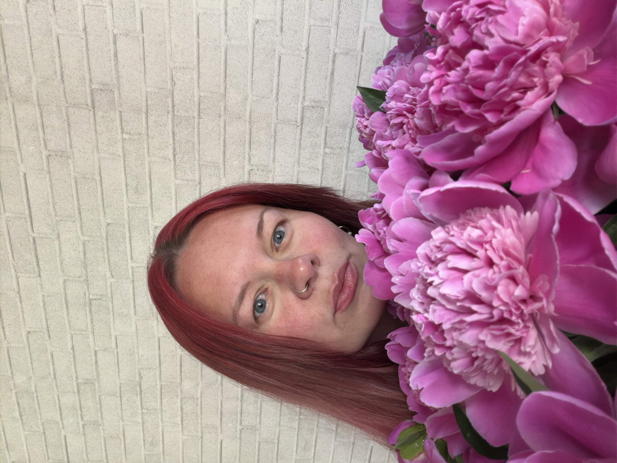The image shows a fair-skinned woman with red hair, a nose ring, and blue eyes, with a large bouquet of pink peonies partially obscuring her face. She is set against a light-colored, textured brick wall. The location and city are unknown.