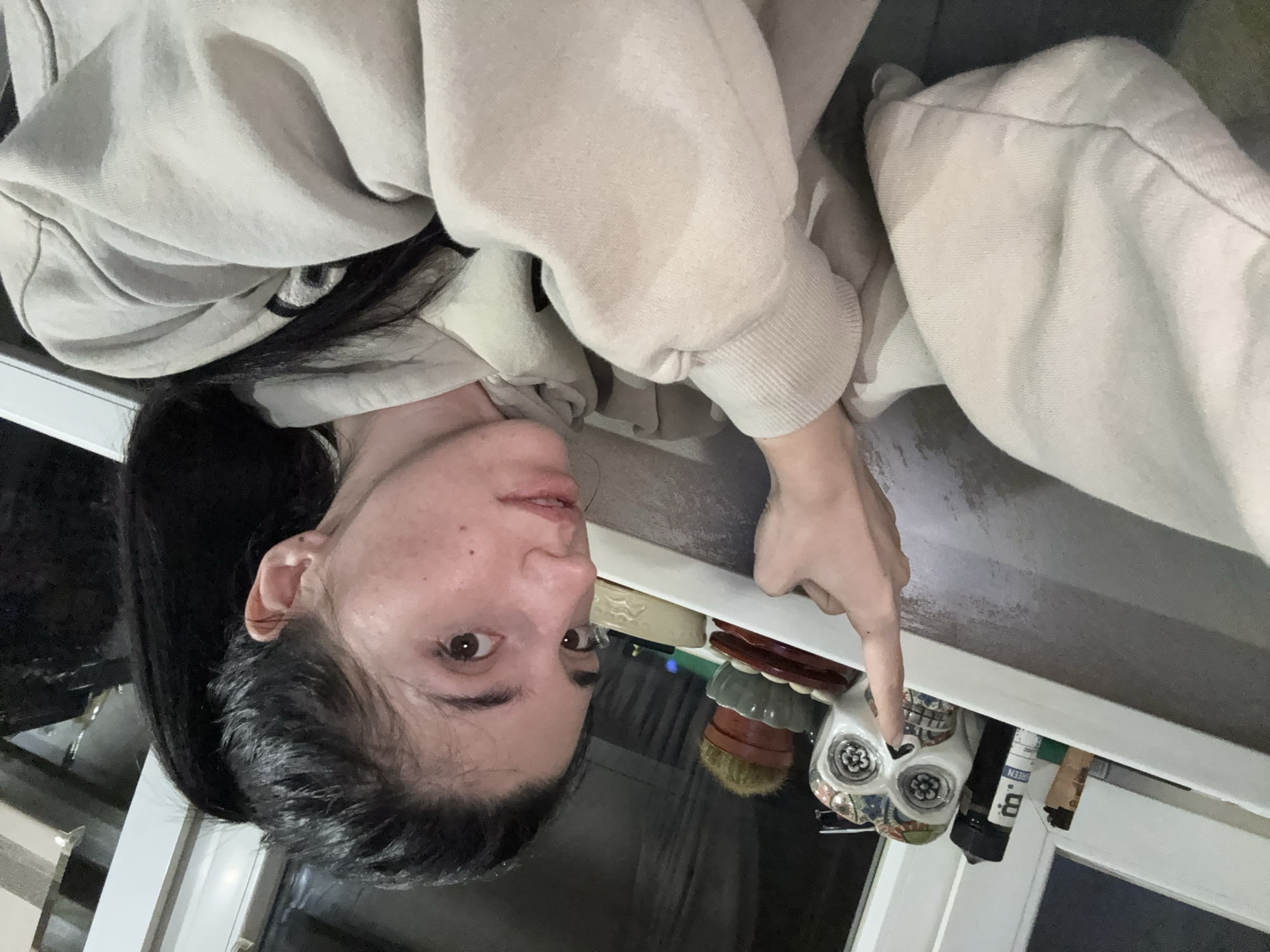 A woman with dark hair in a ponytail, wearing a light beige sweatshirt, is pointing with her right index finger at a decorative skull placed on a shelf. The shelf is also holding other items, including several small jars, a soap bar, and other containers. A window is visible below the shelf. The location in Sofia, Bulgaria, is not discernible from the visual information.