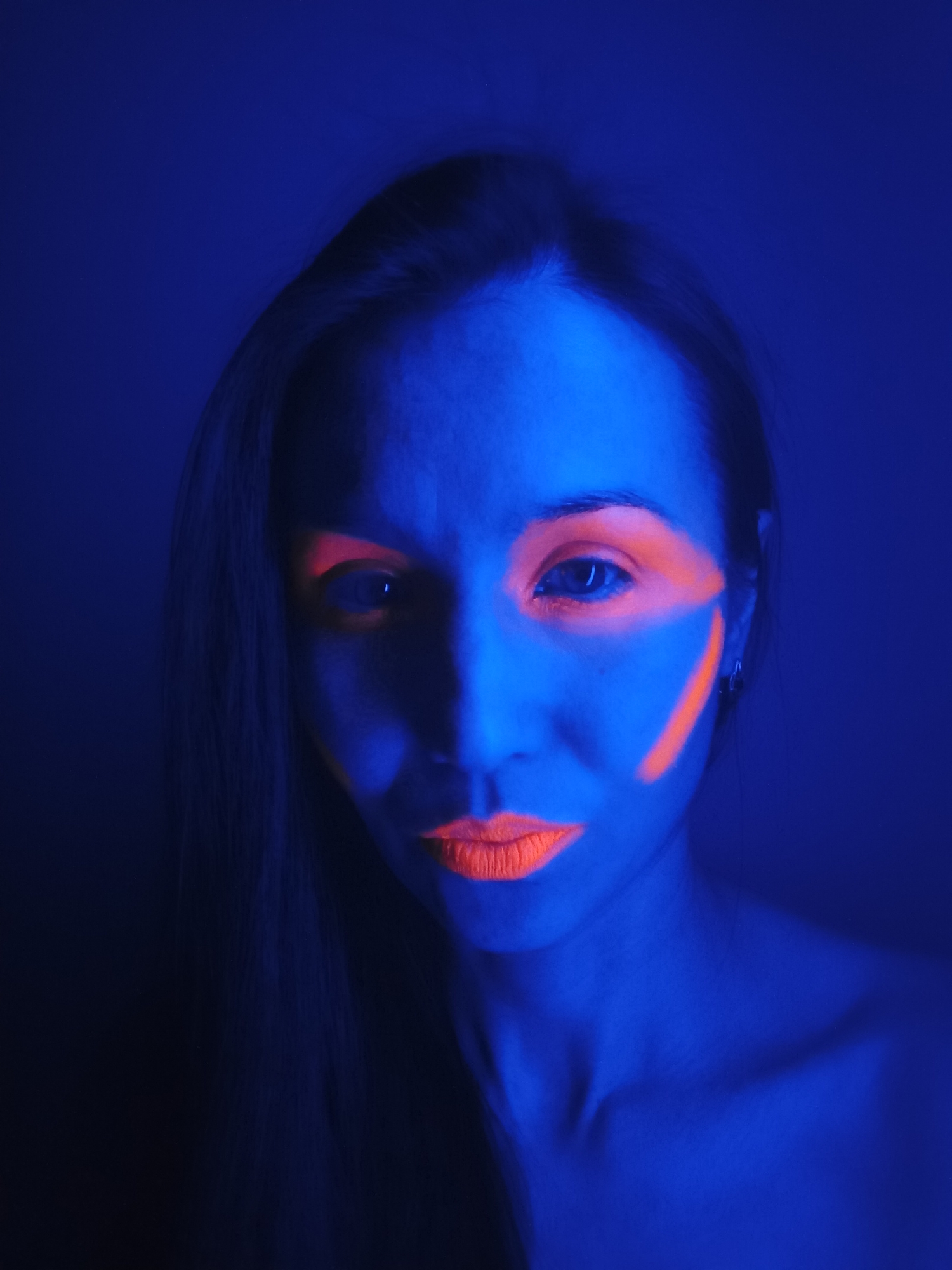 A close-up, artistically lit portrait captures a woman illuminated by ultraviolet (UV) light, rendering her skin and the background in deep shades of blue and purple. Her long, dark hair is visible on the left side of the frame, falling over her shoulder.
The woman wears striking, neon orange, UV-reactive makeup. A prominent, angular design highlights her right eye (viewer's left), extending from the inner corner, above the eyebrow, and towards her temple. A separate diagonal streak of the same glowing orange is painted on her right cheekbone. Her lips are also coated in the vibrant, fluorescent orange lipstick, creating a strong contrast with the blue light.
She looks directly at the camera with a neutral and focused expression. Her bare shoulders and clavicle are visible, suggesting an intimate or studio setting. The overall mood is artistic, futuristic, and dramatic, emphasizing the interplay of the intense blue light and the glowing orange elements. No text is visible in the image. The location context provided is Pervomaysky, Russia.