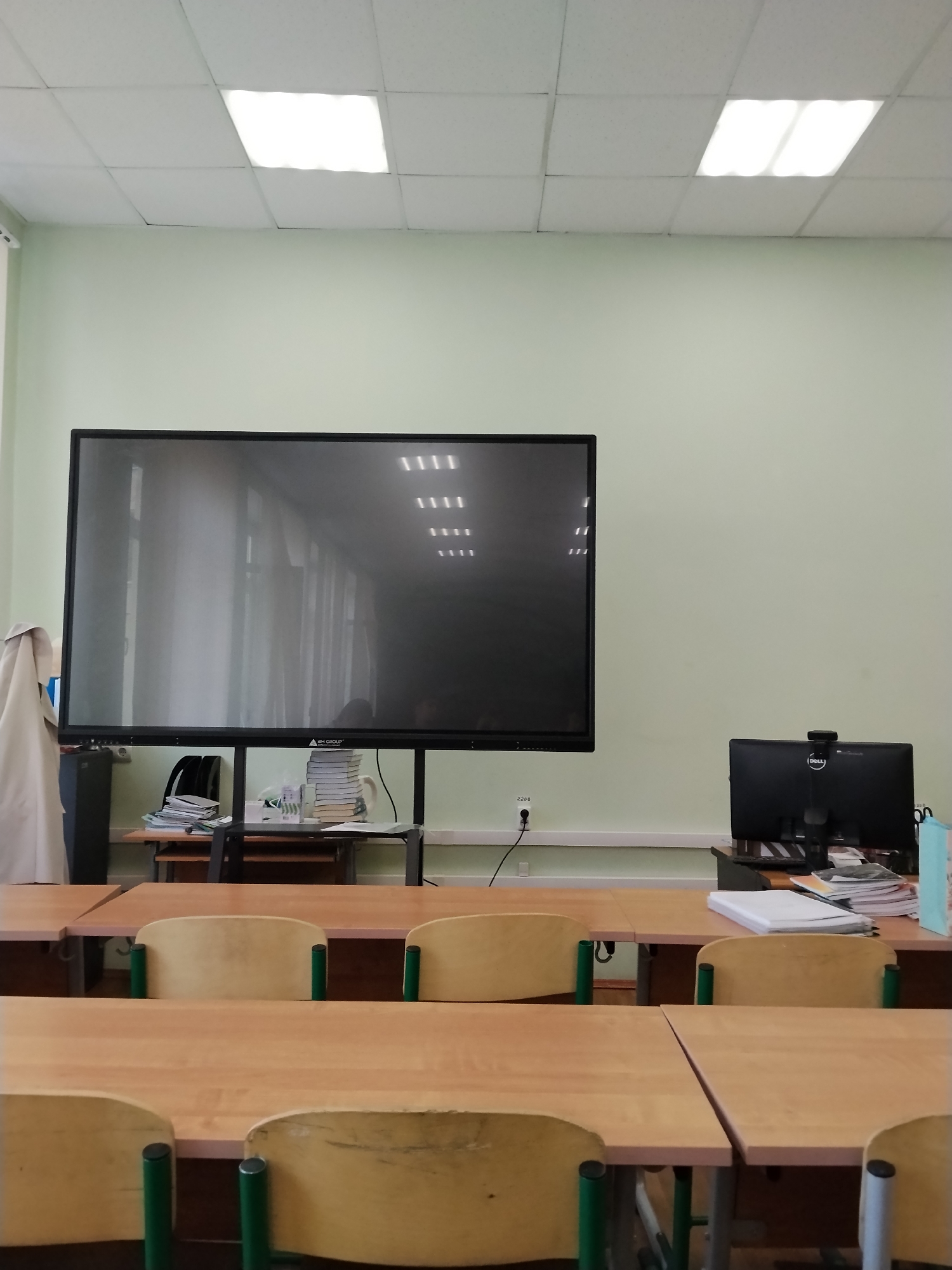 The image depicts a classroom setting in Moscow, Russia, as suggested by the interior design and the specific models of equipment visible.
Environment: The room is enclosed, with a light-colored, possibly pale green, wall paint. The ceiling features recessed fluorescent lighting. The room appears to be well-lit.
Objects: The classroom is furnished with student desks in the foreground, arranged in rows. The desks are wooden with green metal supports. In the background, there's a large interactive display or smartboard mounted on a stand. Next to it, a smaller desk holds a Dell desktop computer with a webcam attached. There are also stacks of books and papers on the desk, along with a few other miscellaneous items.
People: The image doesn't directly show any people, but the setting suggests a space intended for teaching and learning.
Actions/Interactions: Without people present, the scene is static. However, the presence of the desks and teaching equipment implies the potential for interaction, teaching, and studying.
Additional details:
- There's an electrical outlet visible near the smartboard.
- A coat or garment is hanging on the left side, partially obscuring the smartboard's stand.