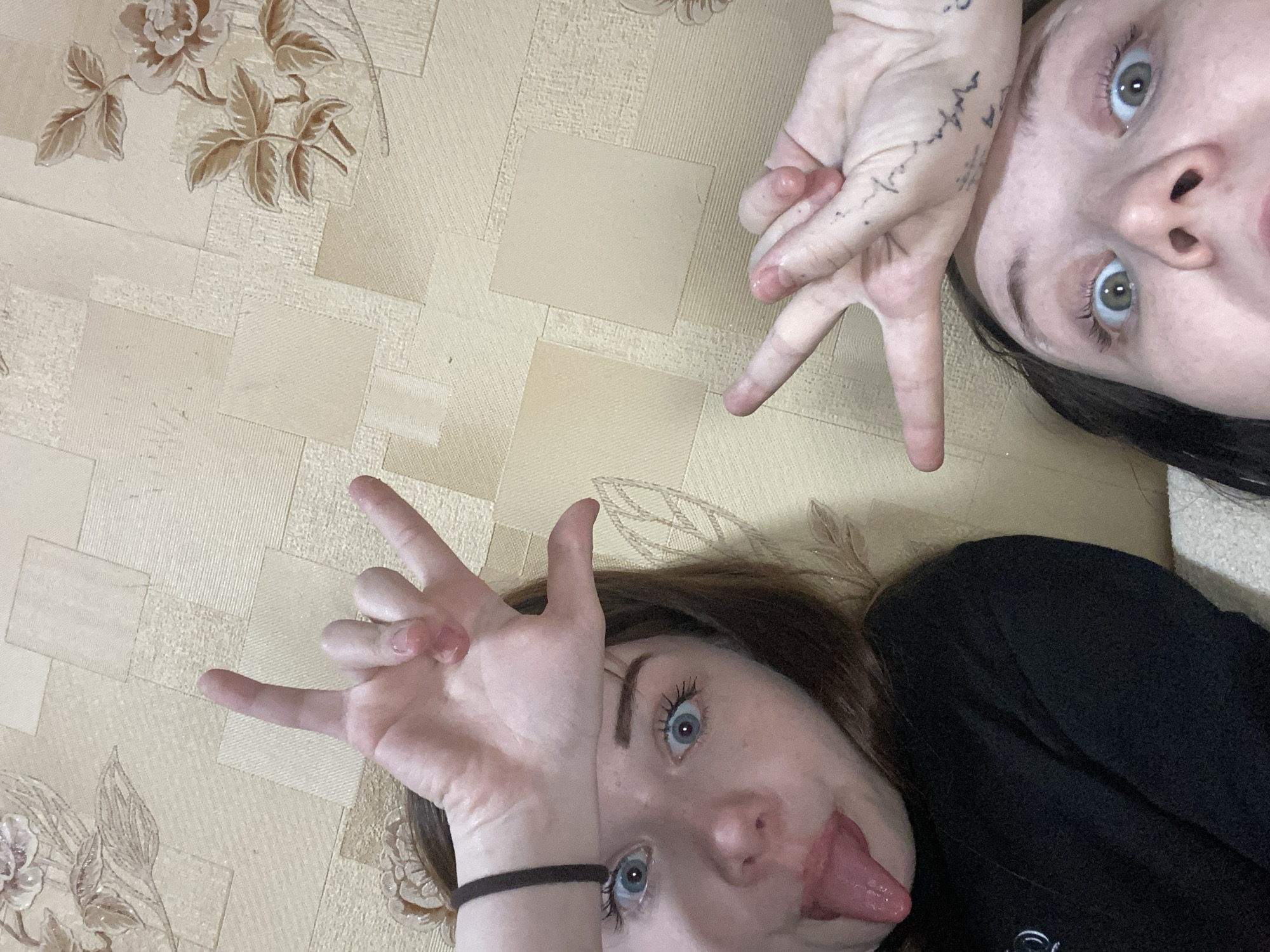Two young women are posing for a selfie on what appears to be a patterned floor. The woman on the right has dark hair, light eyes, and a tattoo on her hand, forming a "peace" sign. The woman on the left has light eyes and is sticking her tongue out while making hand gestures. The location is identified as Chaikovsky, Russia, though there are no specific landmarks or cues to confirm this. The setting is domestic, indicated by the floor covering.