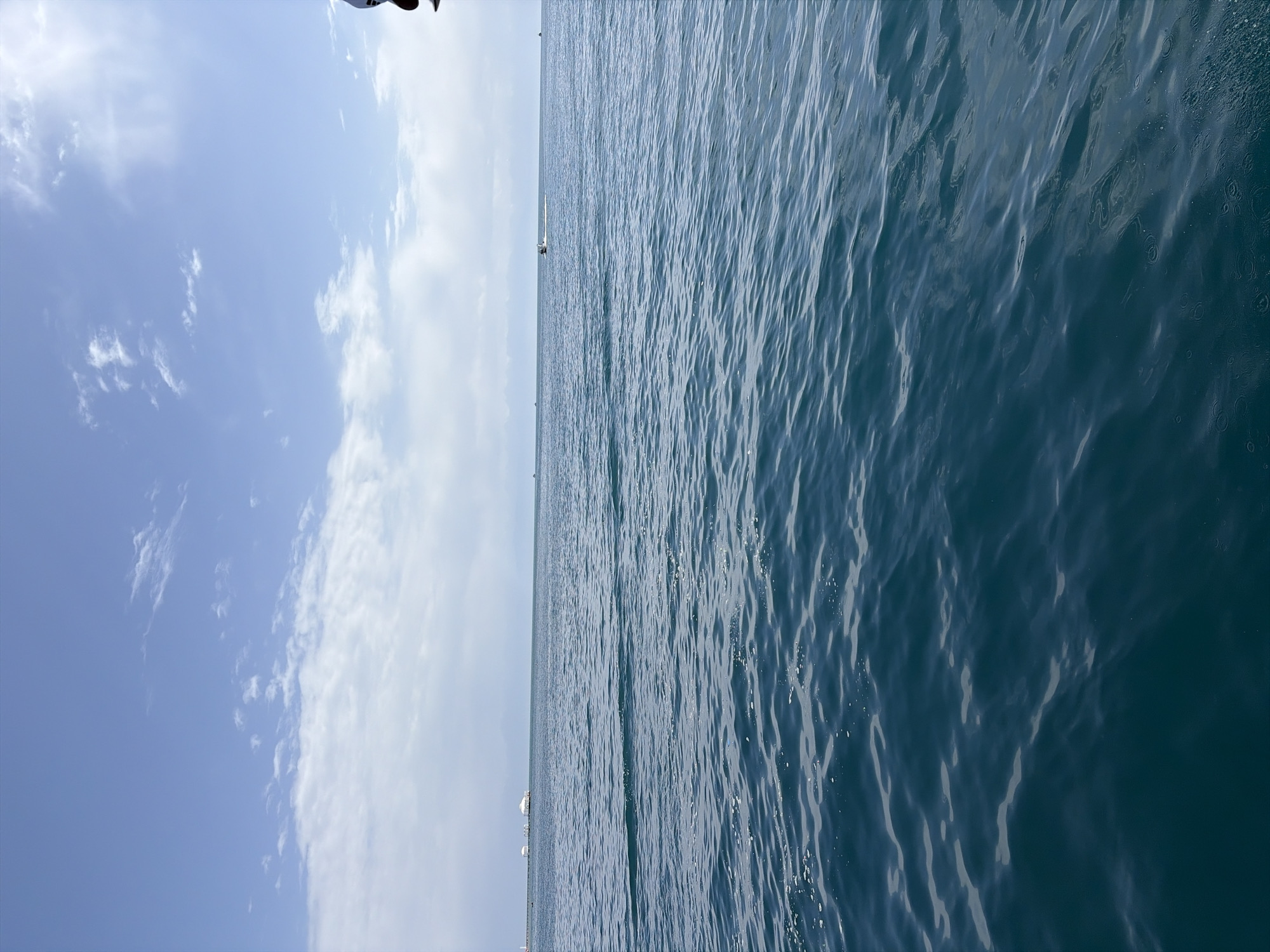 A wide shot captures the serene expanse of Lake Issyk-Kul in Cholpon-Ata, Kyrgyzstan. The deep blue water fills the foreground and midground, its surface gently rippled by a light breeze. In the distance, a hazy horizon separates the water from a pale blue sky dotted with scattered white clouds. A small, distant boat can be seen moving across the water, leaving a subtle white wake. On the far left of the horizon, faint structures suggestive of buildings or piers are visible. The lighting suggests it is daytime, with diffused sunlight creating a soft illumination across the scene. The overall atmosphere is peaceful and open.