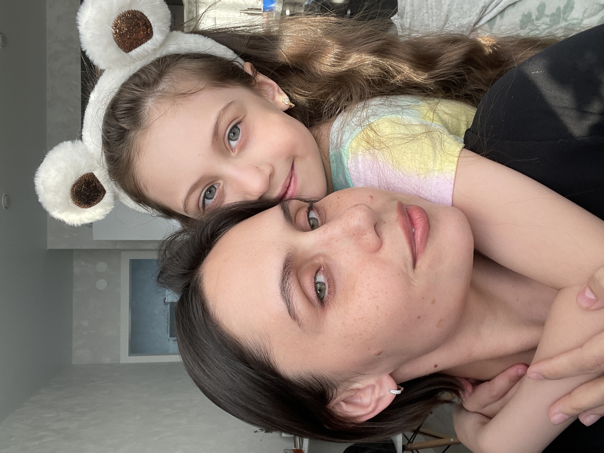 A woman and a young girl are positioned closely together, posing for a picture. The girl, who is wearing a pair of fuzzy bear ears on a headband, has long brown hair. The woman has short, dark hair. They are in what appears to be an indoor setting, with a partially visible window and neutral-colored walls in the background. The exact location in Izhevsk, Russia, cannot be determined from this image.