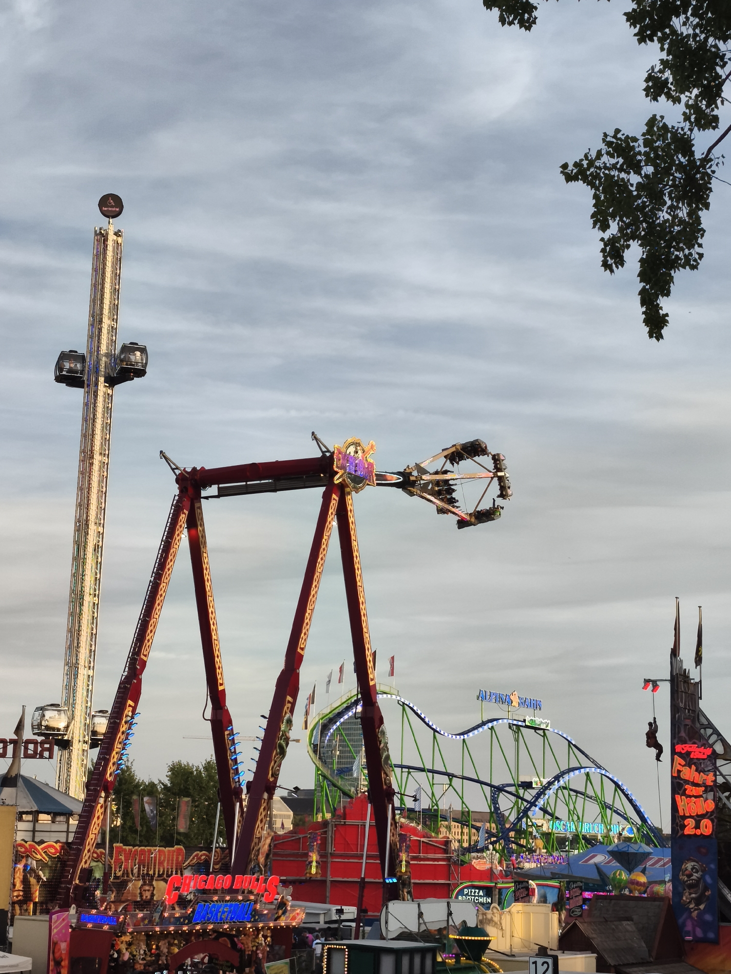 The image captures a bustling scene at the "Größte Kirmes am Rhein" (Largest Funfair on the Rhine) in Düsseldorf, Germany. In the foreground, a large, red amusement ride with yellow accents and flashing blue lights is prominently displayed. This ride features a central arm with suspended gondolas where individuals are seated. To the left, a towering amusement ride, possibly a drop tower, stands tall against the cloudy sky, with two passenger pods visible at its summit. In the middle ground, a green and blue roller coaster with illuminated tracks curves through the air, with a train of cars visible on its structure. Various other amusement park structures, stalls, and attractions are visible throughout the scene, including illuminated signs with German text such as "ALPEN ZIEGE," "SCHICHT," and "FAHRT ZUR HÖLLE 2.0." The sky is overcast with a mix of light and dark clouds. A few people are visible in the distance, appearing to be patrons of the funfair. A tree branch with green leaves is seen in the upper right corner, partially obscuring the sky.