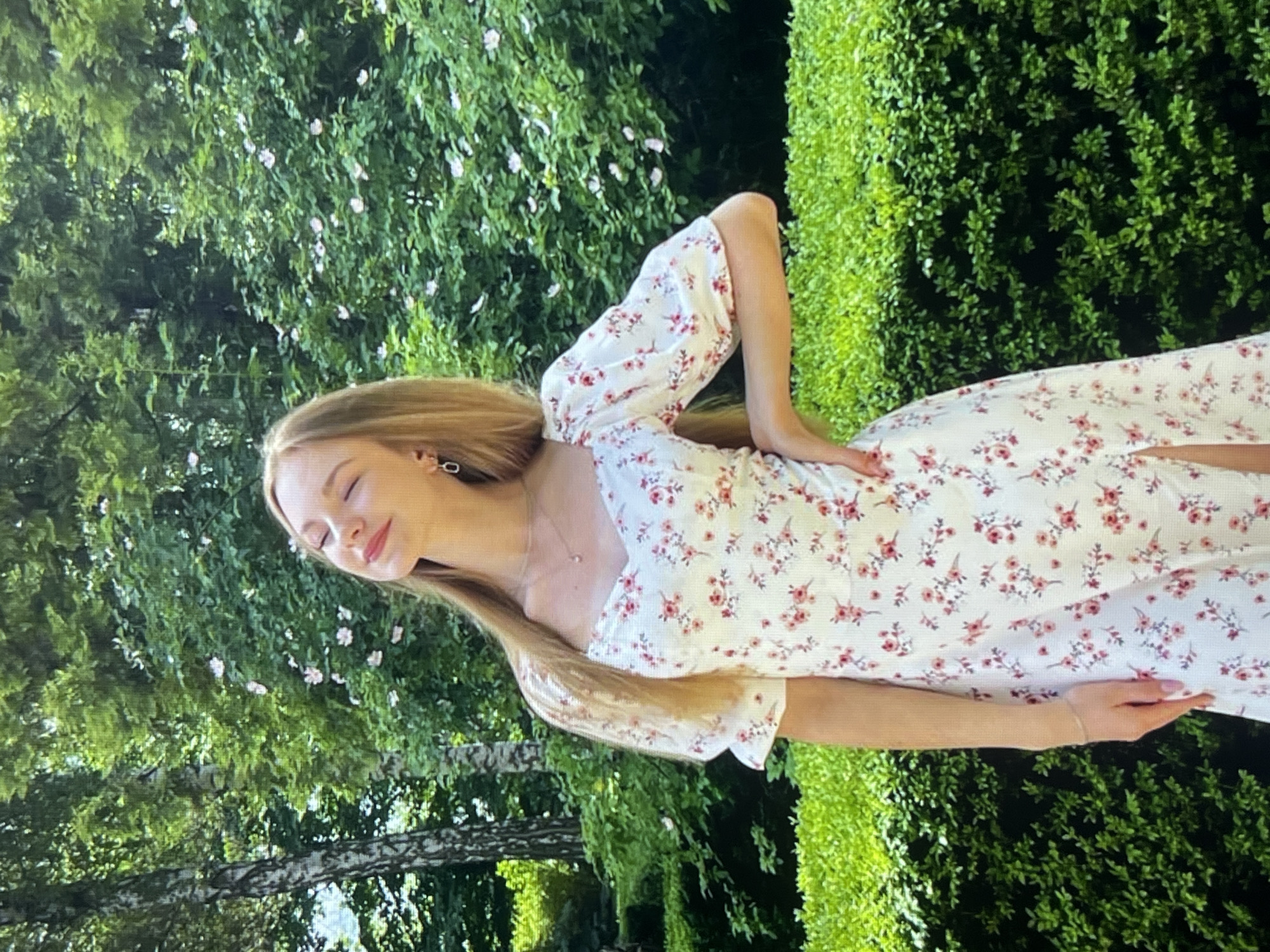 A young woman with long blonde hair and closed eyes stands in a garden. She is wearing a white dress with a floral pattern. Her right hand is on her hip and her left arm is bent at the elbow. The background is filled with lush green foliage and some small pink flowers. There is a hedgerow in the foreground.