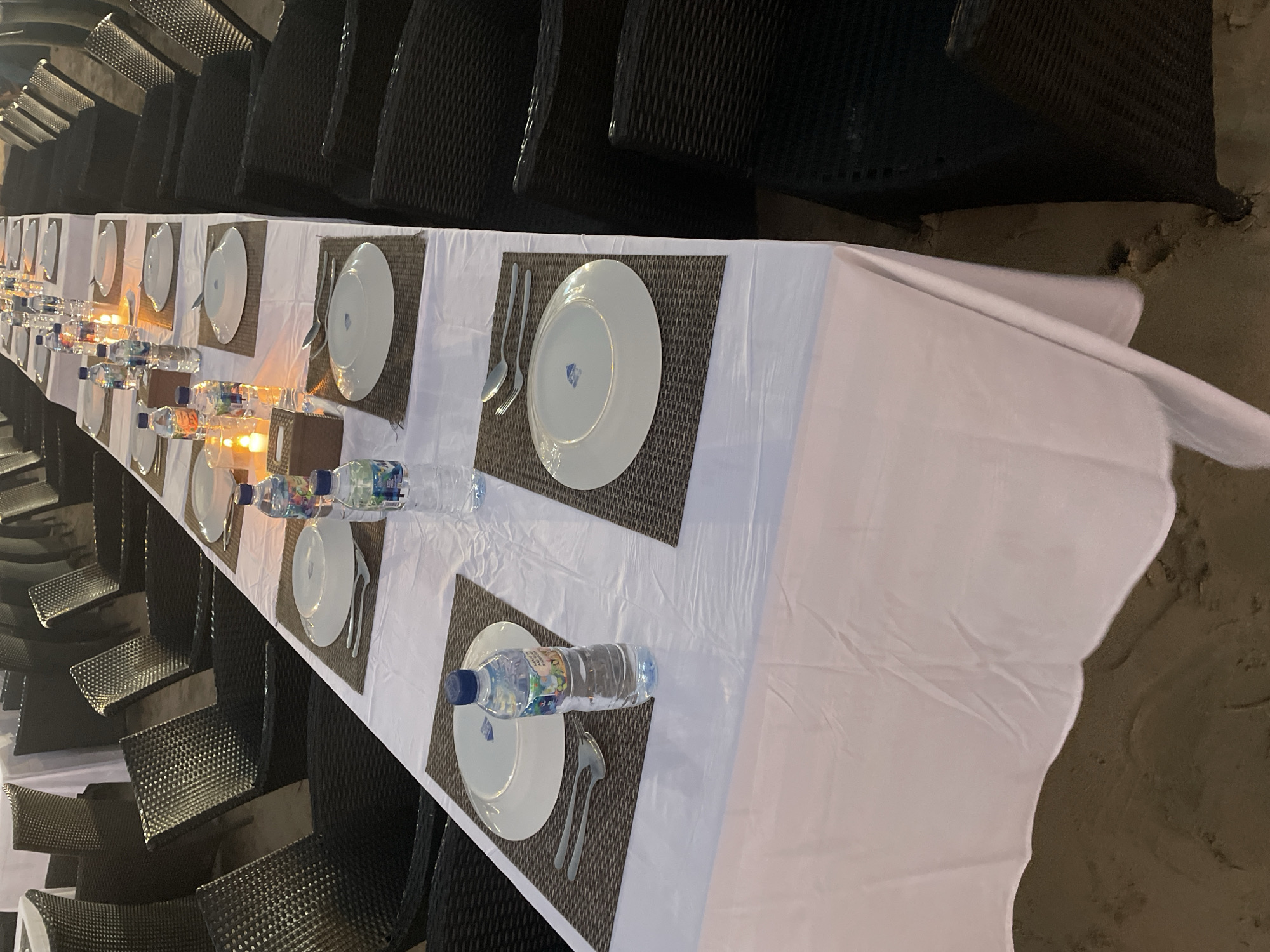 The image shows a long, set table with chairs on either side, suggesting an outdoor dining event. The table is covered with a white tablecloth and set with plates, silverware, and bottles of water. There are also candles on the table, casting a warm glow. The chairs appear to be made of a dark, woven material. The ground beneath the table appears to be sand, indicating a beach or coastal location. The overall lighting suggests it is evening or nighttime. No people are visible in the image.