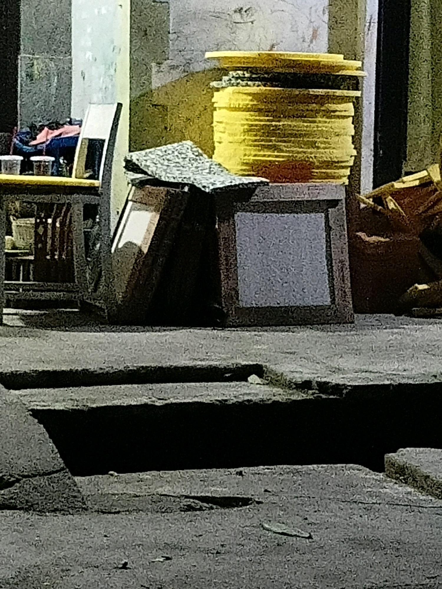 The image shows an outdoor scene with stacked yellow circular objects in the background. In the mid-ground, there is a dark wooden frame with a reflective surface, possibly a mirror or glass, leaning against a darker object. A patterned piece of material is resting on top of the frame. To the left, a low wooden table holds three white containers, and behind that, a piece of blue fabric with pink material on top is visible, possibly clothing. Further left, a dark chair with a light-colored back is also present. The foreground consists of concrete steps and a textured ground. The scene appears to be in a somewhat cluttered outdoor area, possibly a storage space or a sidewalk in the city of Cikal, Indonesia, judging by the context provided. There are no people visible in the image.