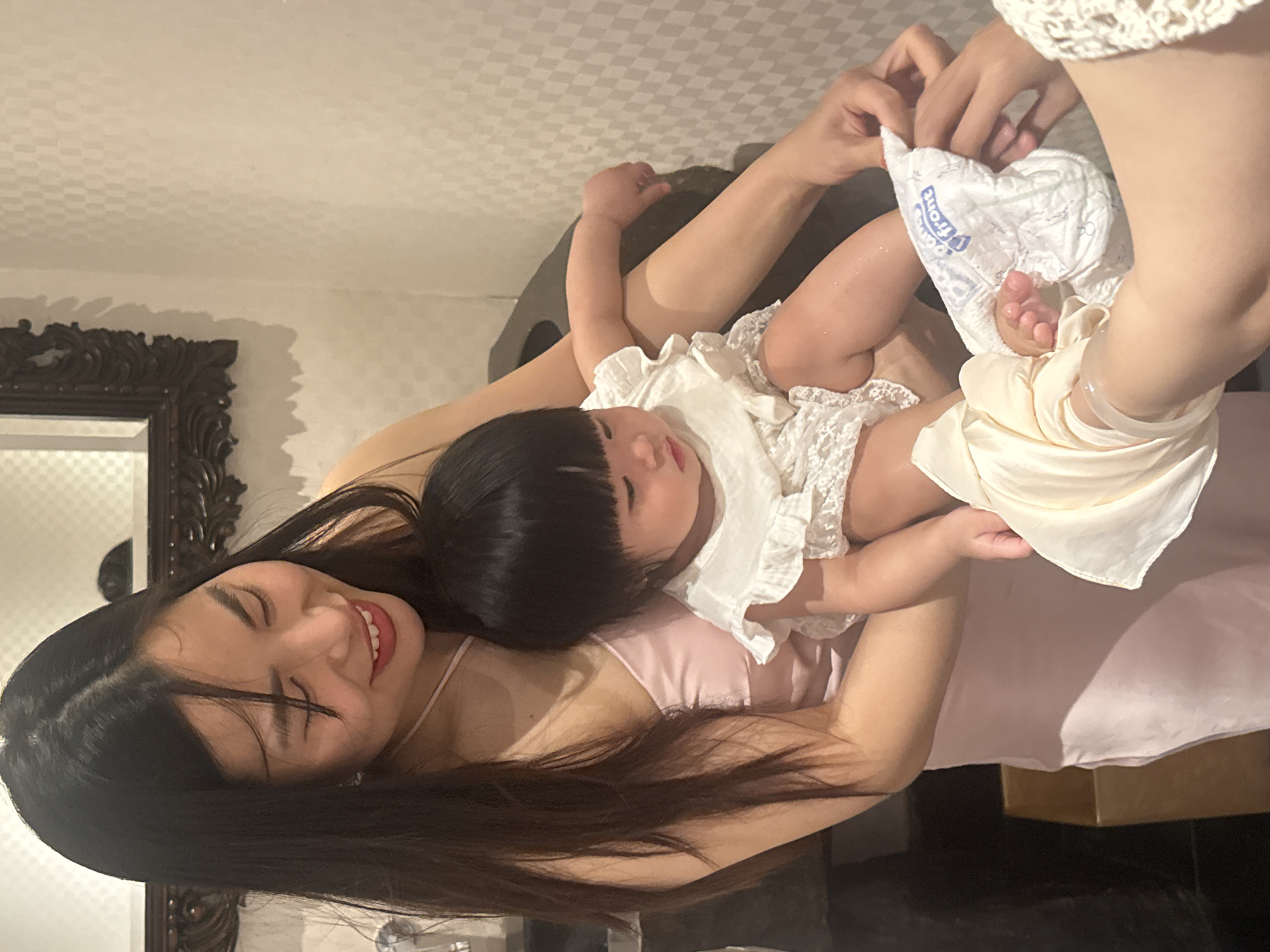 An indoor scene depicts three individuals engaged in an interaction. A long-haired adult female, wearing a light pink spaghetti-strap dress, cradles an infant with dark hair and bangs, dressed in a white ruffled and lacy romper. The adult female exhibits a soft smile while looking down at the infant. The infant's gaze is directed downwards towards its feet. A second individual, whose hands and forearms are visible from the right, is performing an action on the infant's lower body. One hand, partially covered by a cream-colored garment or glove, holds the infant's right foot. The other hand is near the infant's left leg, where a white disposable diaper, partially marked "Front" in blue, is visible. The immediate background features a light-colored wall with a repeating checkered or square pattern. To the left, a dark, ornate-framed mirror is mounted on the wall. A dark, possibly upholstered surface is partially visible behind the infant. The setting appears to be an upscale interior space, possibly a restroom, changing room, or spa treatment area. The specific location within Đà Nẵng, Vietnam, is not discernible from the image.