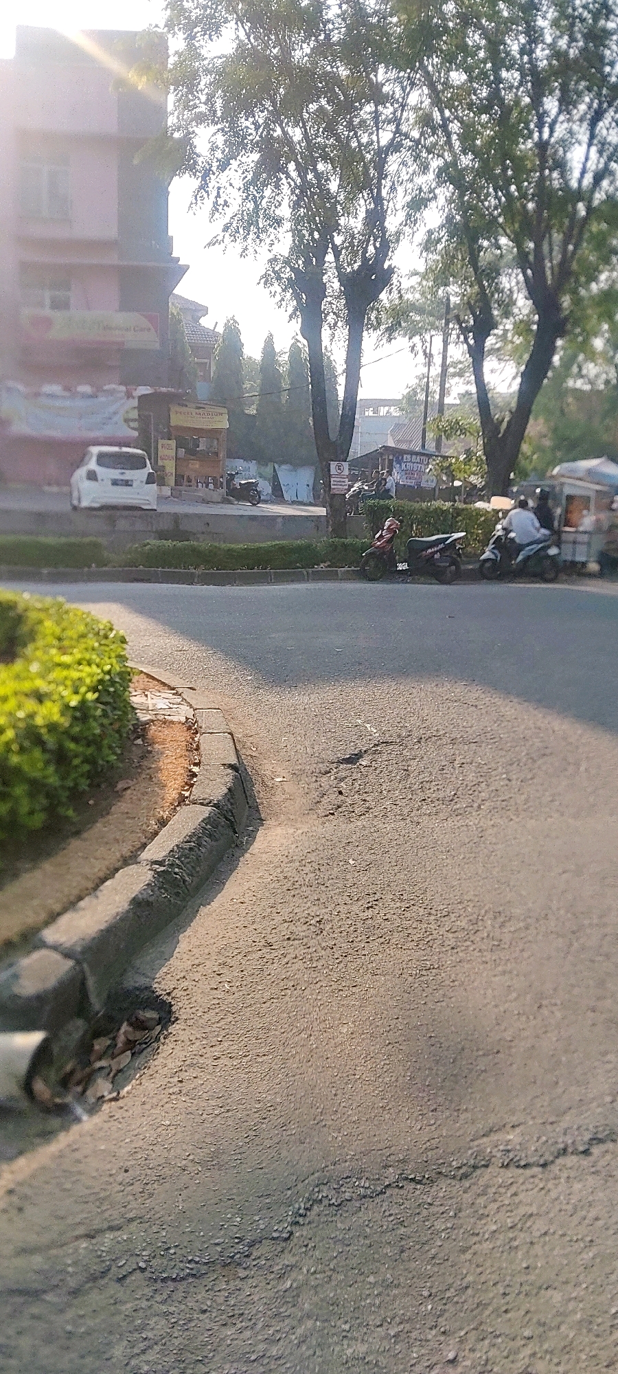 The image captures a street scene in Cibatu, Indonesia, under the warm glow of morning sunlight. In the foreground, the rough asphalt road takes up a significant portion of the frame, showing signs of wear and tear with cracks and potholes. A concrete curb lines the left side of the road, with a patch of vibrant green foliage growing along its edge, interspersed with dry leaves and debris.
Beyond the curb, a white car is parked on the side of the road, facing away from the viewer. Adjacent to it, a small food stall with a wooden structure is visible. Further down the street, several motorcycles are parked, and a few people are gathered around another food stall. One person is seen riding a motorcycle, and another person, possibly a passenger, is on the back of a separate motorcycle.
In the background, large trees with dense foliage cast shadows on the road. A multi-story building, painted in a soft pink hue, stands on the left side of the image, with visible windows and balconies. The sky is bright, suggesting a clear day. The overall impression is that of a bustling urban environment in the early morning, with daily activities beginning to unfold.