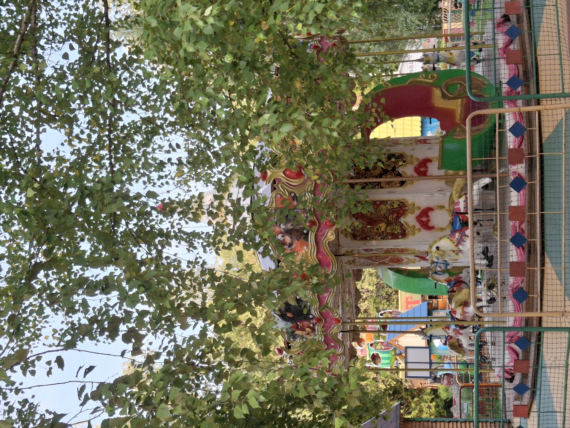 A carousel, featuring painted horses and a decorative top, is centered in the frame, partially obscured by green foliage from overhead trees. The carousel is located in an amusement park in Chaikovsky, Russia, evidenced by a sign in the background that reads "ВЫХОД" (Exit) in Russian Cyrillic, and an adjacent structure with Russian text. The carousel is surrounded by a safety fence. In the background, other amusement park attractions are visible, including colorful structures and rides. The sky is a pale blue with some clouds. The overall scene depicts a recreational area with a children's amusement ride.