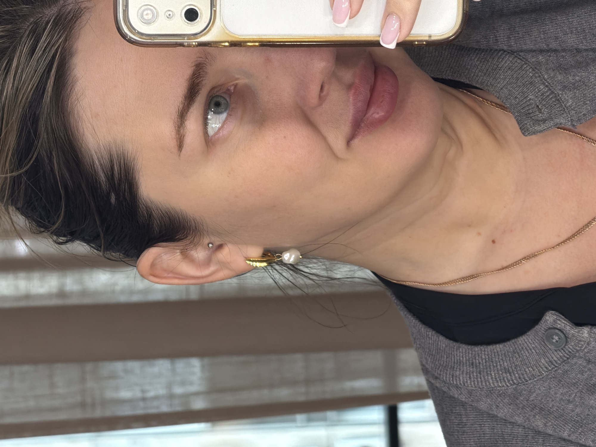 The image is a close-up self-portrait of a woman with light skin, blue eyes, and dark hair. The woman is holding a cell phone, which is positioned so that the camera is near her forehead. The woman has a small piercing in the upper part of her ear, and she is wearing a gold necklace and earrings, with a gold and pearl earring. She is wearing a gray sweater over a black top. The picture appears to have been taken indoors near a window. The location in the city of Khimki, Russia, cannot be determined from this image.