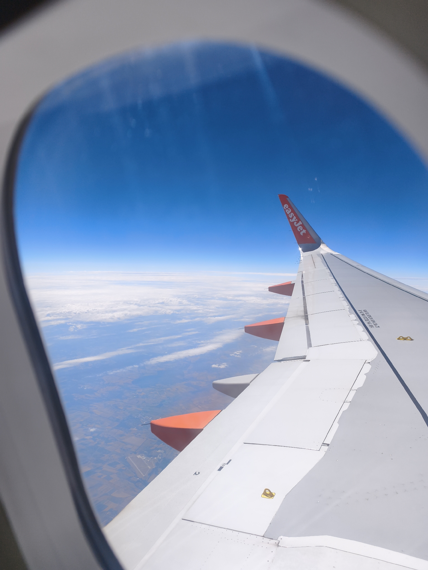 An aerial view of a clear blue sky above a layer of white clouds is visible through an airplane window. Below the clouds, patchwork fields and a small industrial area are discernible. The right side of the image is dominated by the wing of an EasyJet airplane, featuring its distinctive orange wingtip accents. A portion of the airplane's fuselage is also visible, along with the window frame of the aircraft. The wing is made of white material with visible panel lines and some markings. A yellow symbol is present on the wing's surface. The overall scene captures a moment of flight.