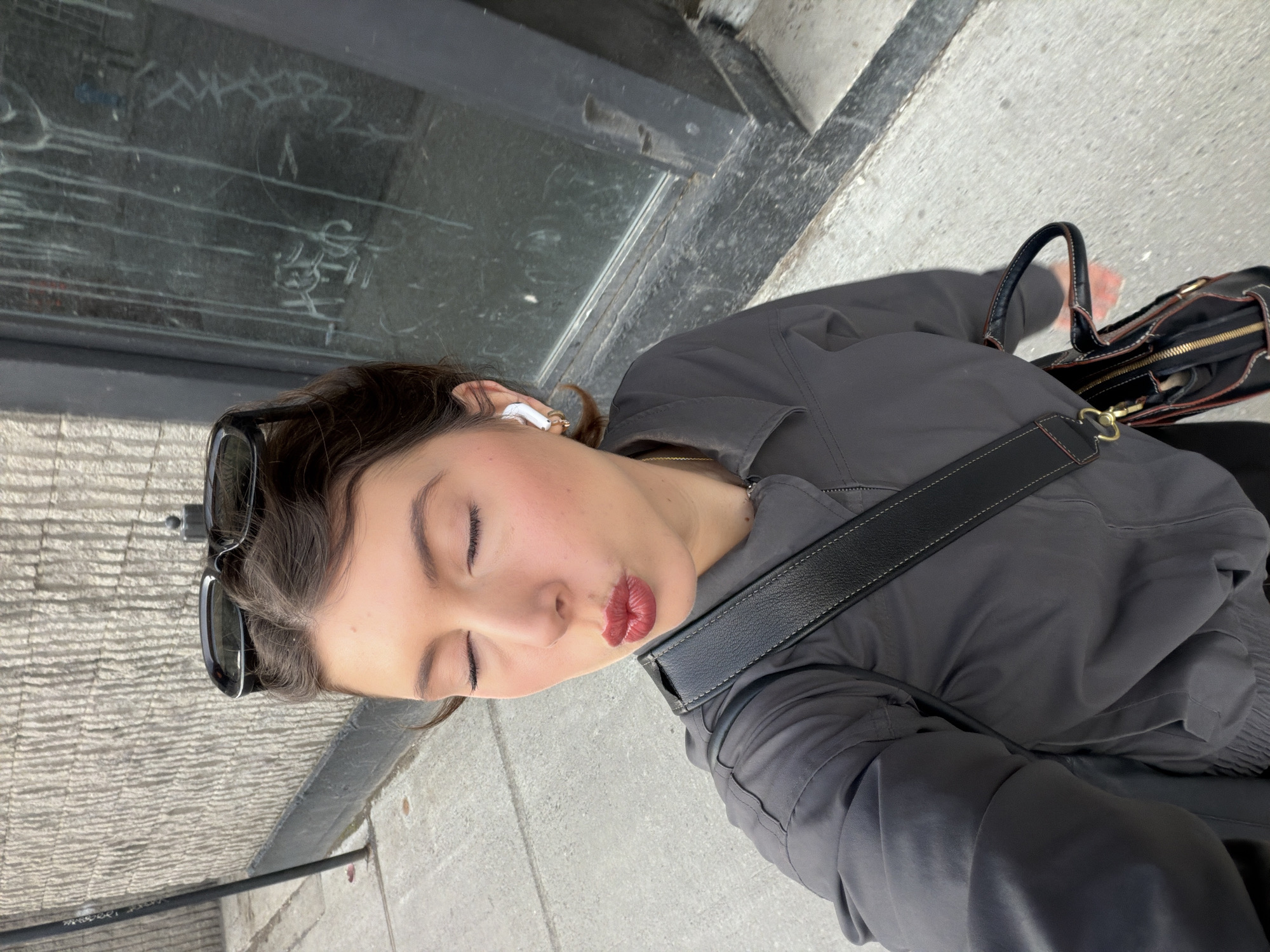A woman stands on a Montreal sidewalk, angled towards a building wall and window with graffiti. She wears sunglasses atop her head, wireless earbuds, a grey jacket, and a purse with a black strap. She is puckering her lips as if to kiss. The sidewalk is grey concrete.