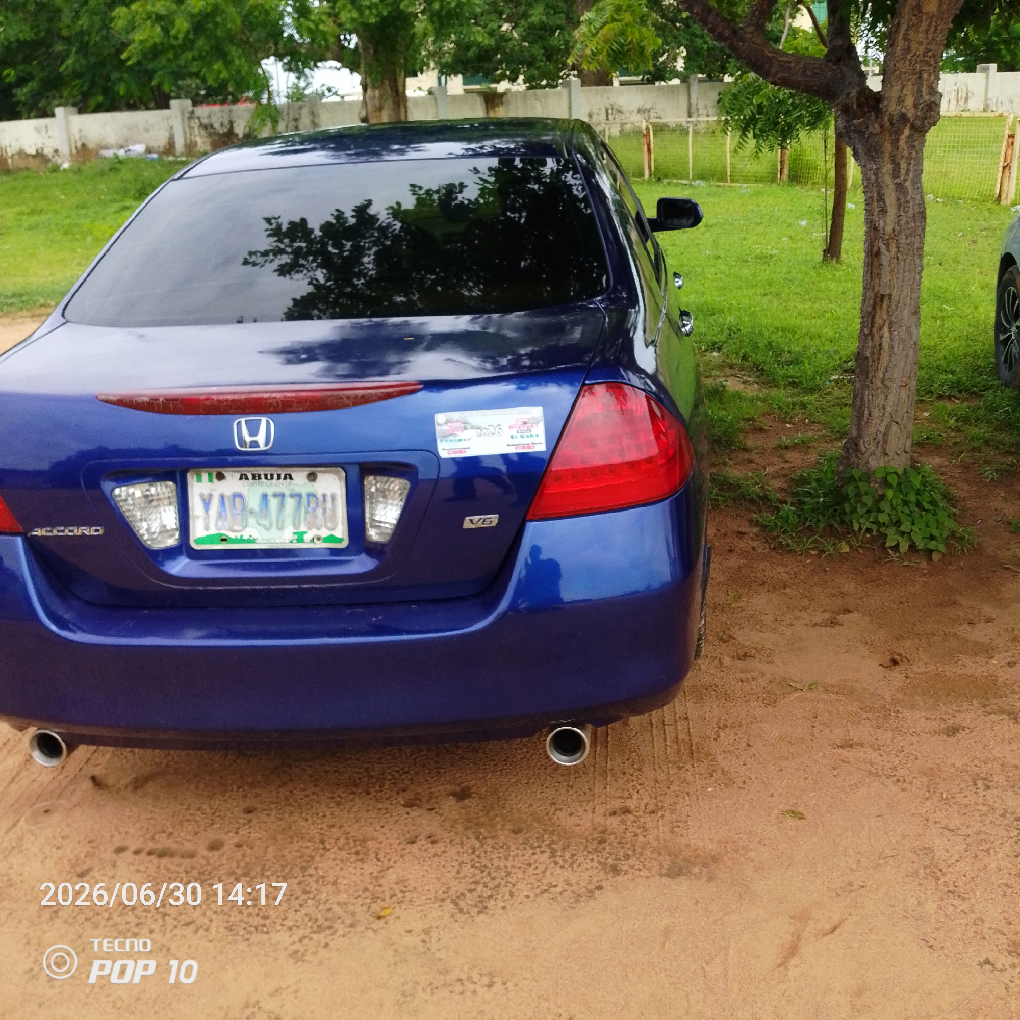 A blue 2007 Honda Accord is parked on a dirt surface in Jalingo, Nigeria. The car's rear is visible, showing its license plate (YAB-477RU) and "ACCORD" badging. A sticker is affixed to the car's trunk, reading "2026" and "Presper." The car is situated in an outdoor environment with a grassy area, trees, and a concrete wall in the background. The timestamp "2026/06/30 14:17" and "TECNO POP 10" are overlaid on the bottom left of the image.