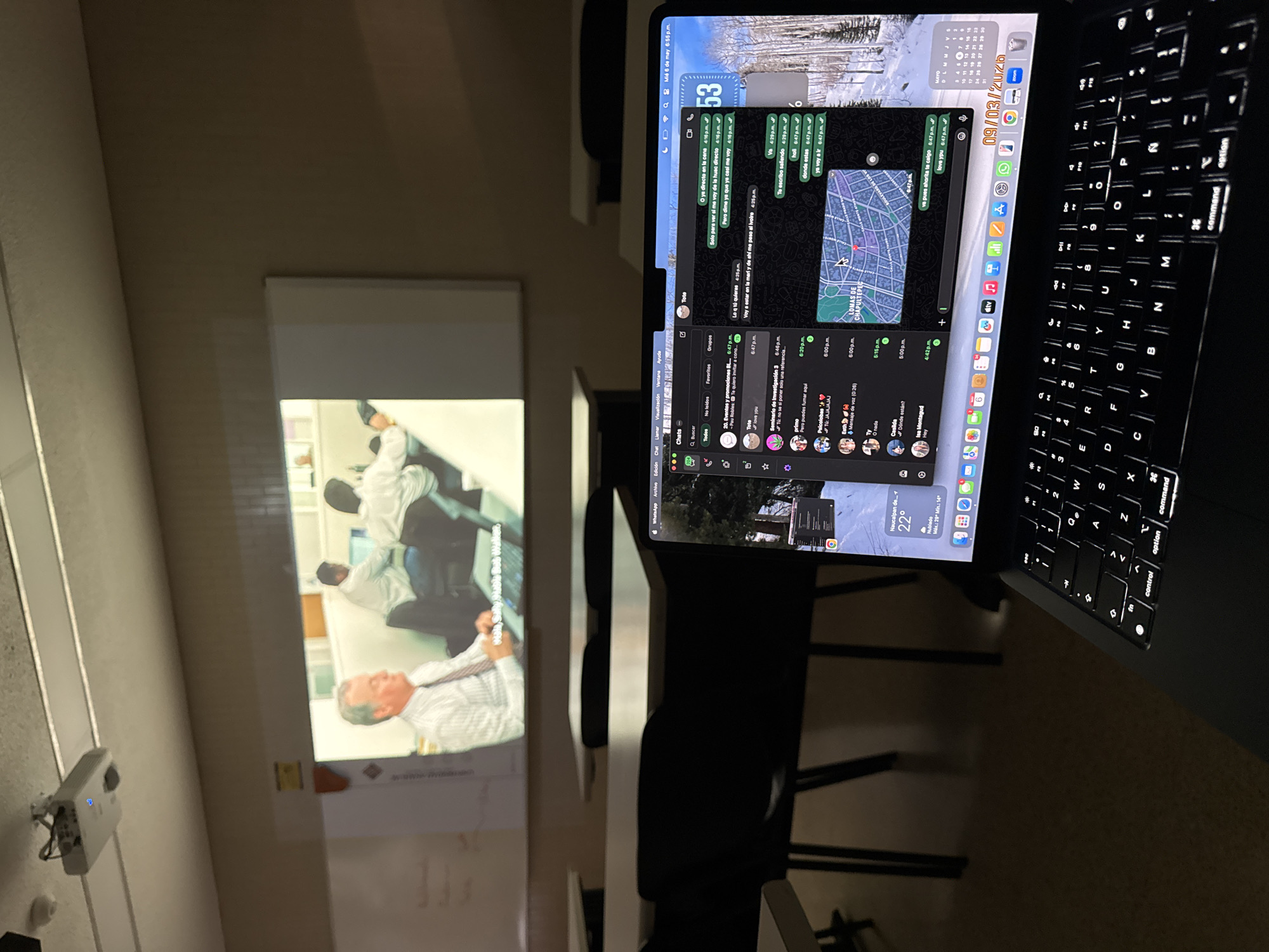 A meeting room with a projector displaying a corporate image on a screen. In the foreground, a laptop displays an open WhatsApp conversation showing the location of a person in Lomas de Chapultepec in Mexico City.