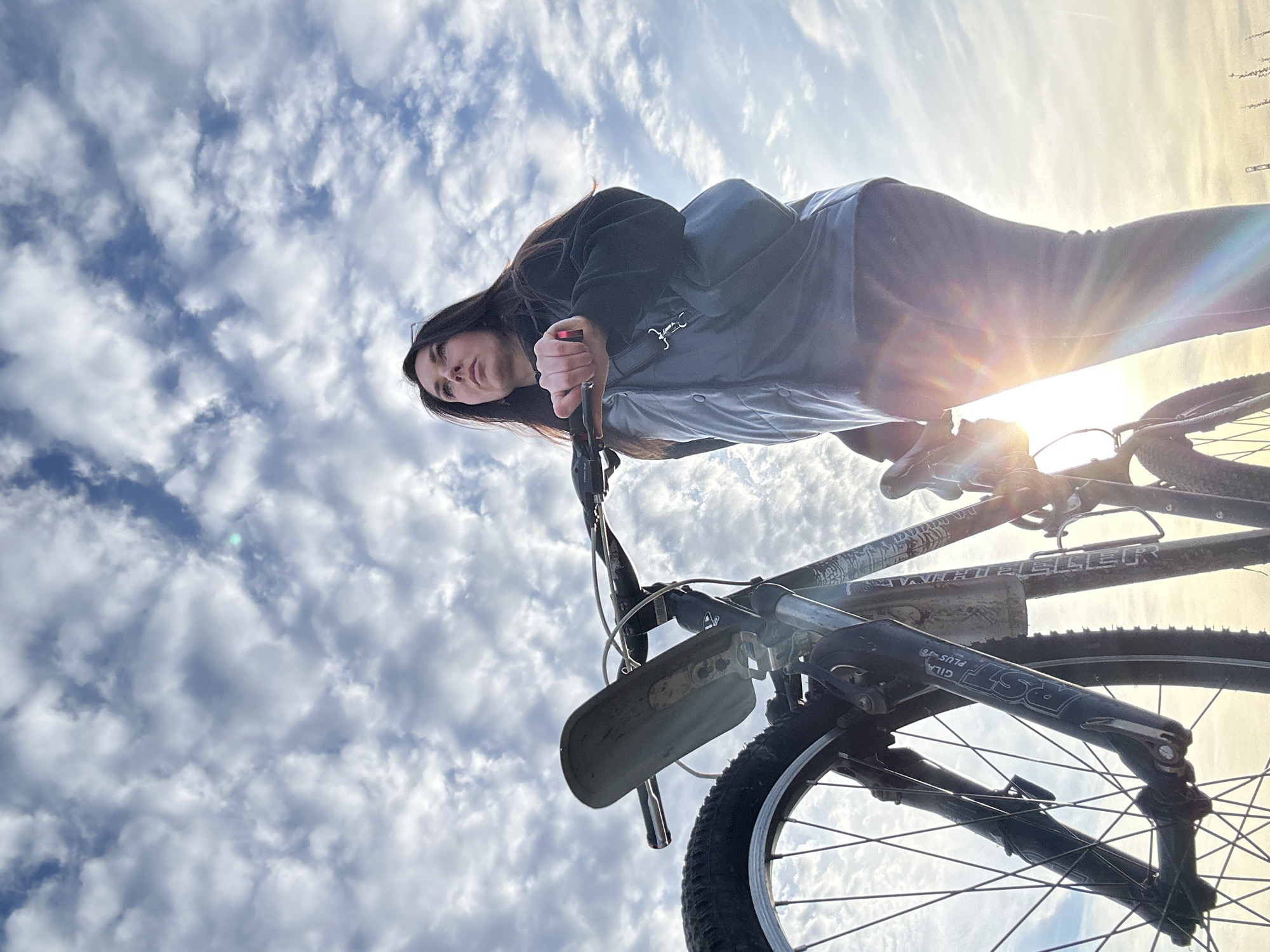 An outdoor, low-angle photograph captures a female subject standing next to a bicycle, observed against a heavily clouded sky in Chaikovsky, Russia. The woman, positioned center-right, has dark hair and looks upwards and to her right. She wears a dark long-sleeved shirt, a grey quilted vest, and dark trousers, with a dark shoulder bag slung across her body. Her right hand grips the right handlebar of a dark-colored mountain bicycle, which occupies the left and central foreground. The bicycle features a dirty, light grey front fender, visible front suspension forks marked "RST Gila Plus T6," and a water bottle cage on its frame. A strong sun flare from the lower right backlights the scene, casting a prominent rainbow effect across the lower right quadrant and illuminating the clouds from behind. The sky is largely covered with white and grey stratocumulus or altocumulus clouds. The ground is minimally visible at the very bottom, appearing as a subtle light-colored horizon line with faint, indistinct structures.