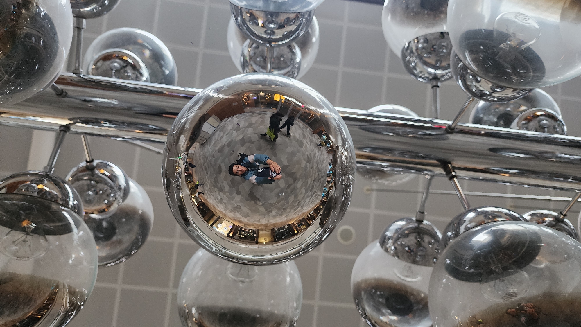 A low-angle shot captures a spherical, mirrored ornament reflecting a person taking a picture of themselves within a large, modern interior space. The floor has a light grey grid pattern. Reflections show storefronts and other people walking. Around the primary sphere are other similar but non-mirrored light fixtures with bulbs visible within. The location is Moscow, Russia.