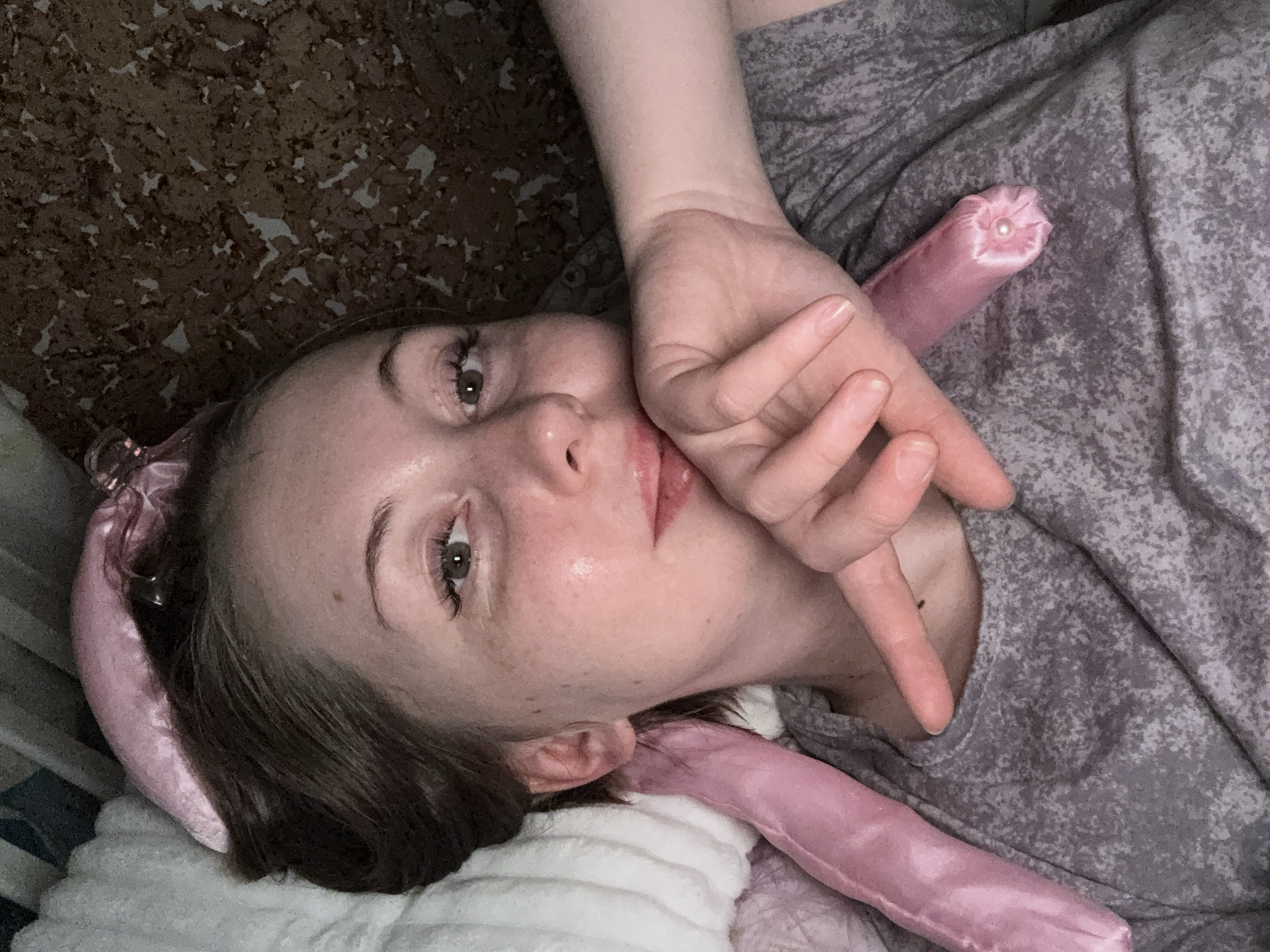A woman lies in bed. She is wearing a gray patterned shirt and a pink silk hair-curling headband. Her left hand is up by her face. The bed is covered with a white blanket. The city is Unknown, Unknown.