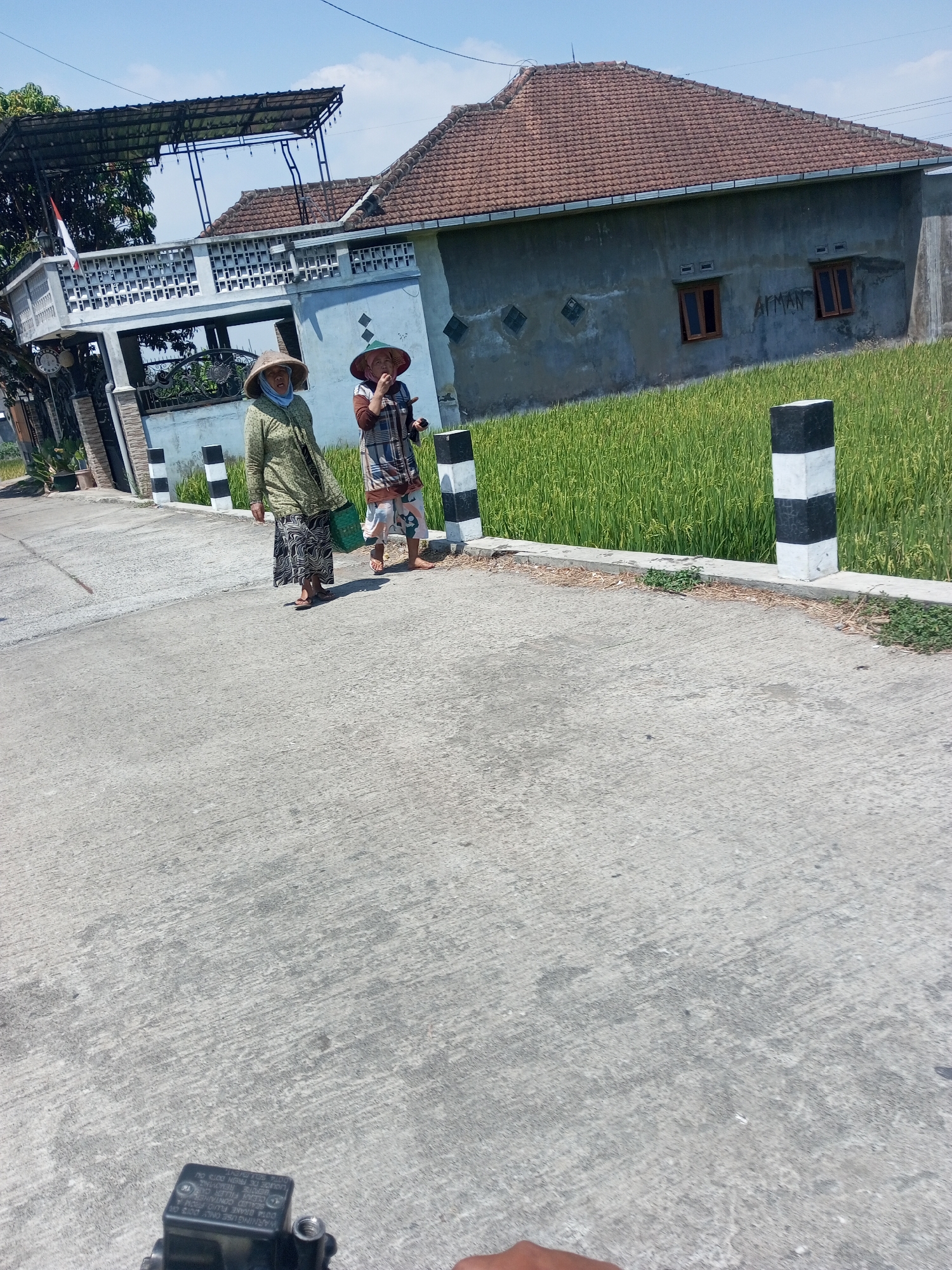 In Purwosari Klampok, Indonesia, on a bright, sunny day, two women walk along a concrete road. Both women wear traditional conical hats, commonly worn by farmers. The woman on the left, dressed in a light green blouse and a patterned skirt, carries a green bag. The woman on the right wears a patterned shirt and a skirt, and appears to be gesturing with her hand. They are walking past a vibrant green rice field that stretches behind a low concrete wall. To the left of the road, a modern house with a tiled roof and two windows is visible, with a decorative white fence in front of it. The scene is framed by clear blue skies and the lush green landscape, suggesting a rural or semi-rural setting. The image appears to be taken from a slightly elevated perspective, possibly from a moving vehicle.