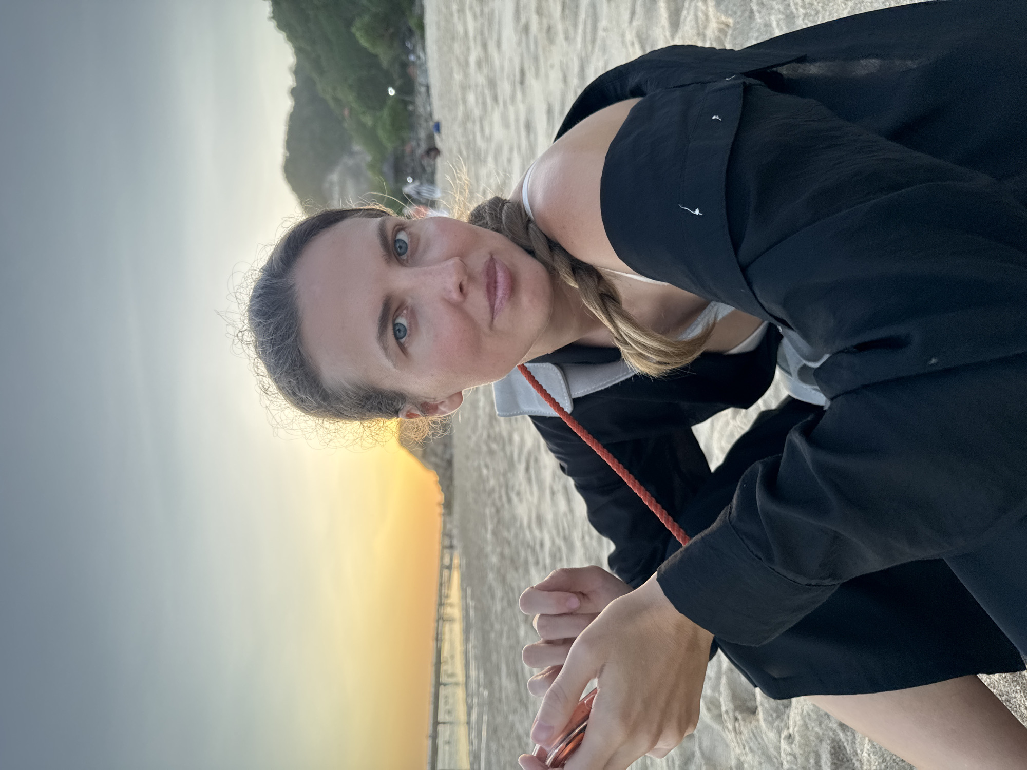 The image features a woman with blonde hair in a braid, wearing a white top and a black open shirt. She is holding a small device or object in her hands. The background suggests a beach environment, with sand and a sunset or sunrise sky. Based on the location, the image was likely taken in Kuta Selatan, Indonesia. A treed hill is visible in the background with buildings.