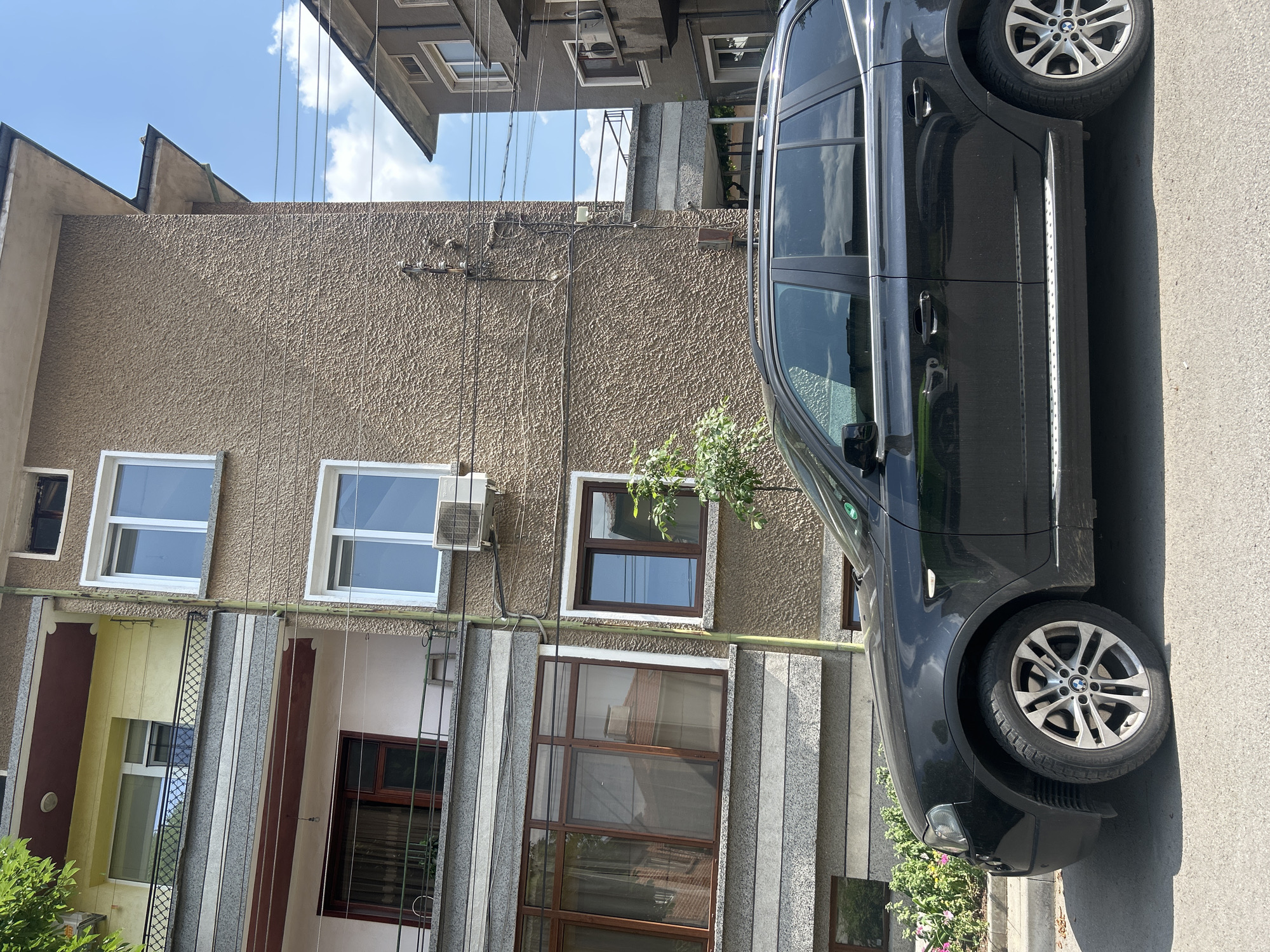 A black SUV, likely a BMW given the visible logo on the wheel caps, is parked on the right side of a paved street, facing left. The car has multi-spoke silver alloy wheels and a silver running board along its side. To the left of the car, a multi-story building with a light brown, textured stucco facade stands.
The building features various windows with white and dark frames, and several air conditioning units are mounted on its exterior. Below the upper story windows, sections of the building facade show different colors and textures, including yellow, maroon, and grey, possibly indicating balconies or recessed architectural features. Numerous black utility wires and cables run across the building's facade and vertically across the scene.
Above the building, a distinctive sawtooth or zig-zagged roofline is visible against a clear blue sky with scattered white clouds. The bright sunlight suggests it is daytime. Some green foliage is visible near the base of the building and the front of the car. The scene depicts a static street view in what appears to be an urban or suburban environment.