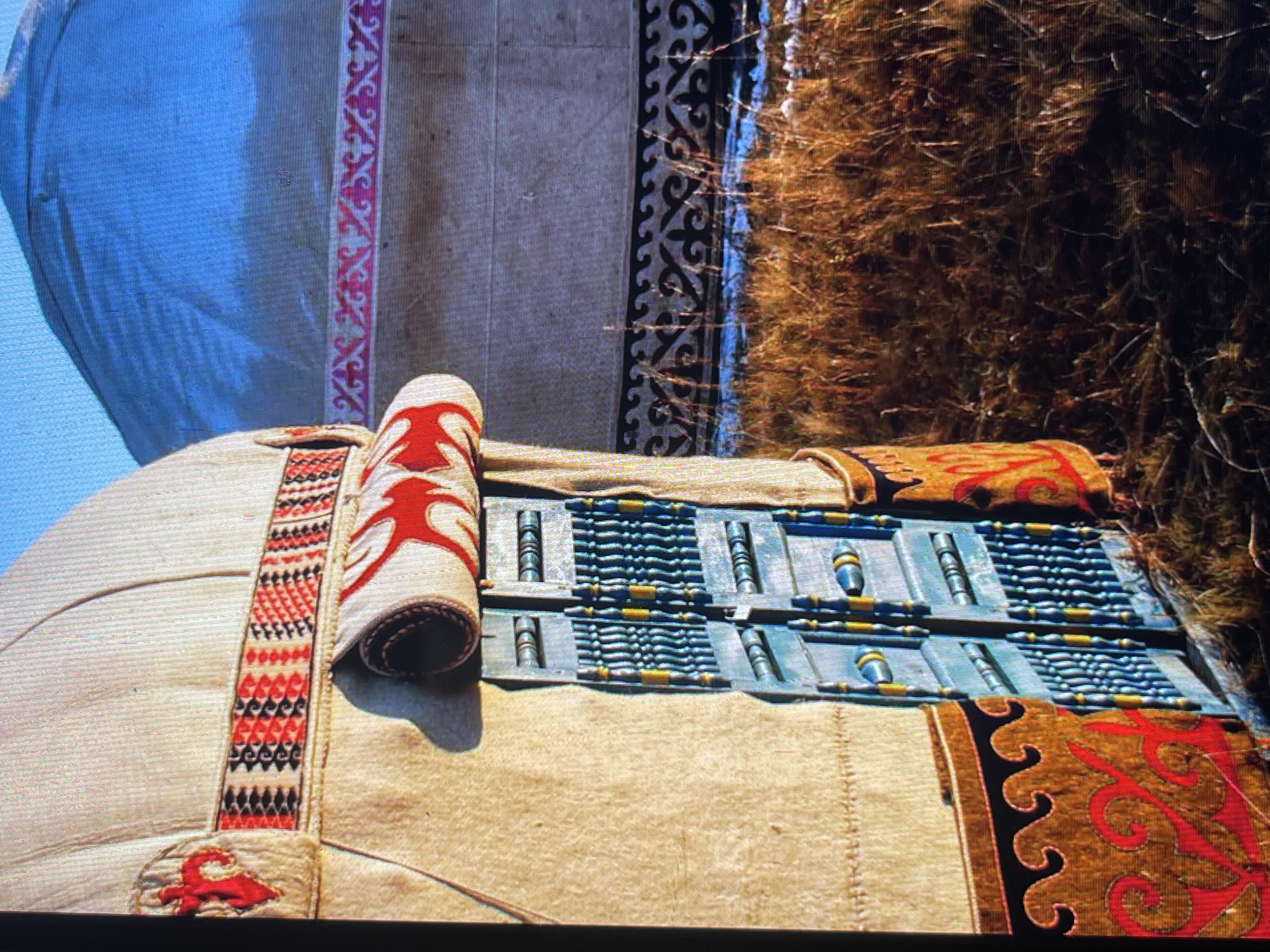 The image shows the entrance of a yurt, a traditional nomadic dwelling, in Bishkek, Kyrgyzstan. The yurt is decorated with colorful woven fabrics and intricate patterns. The door is made of blue wood with yellow accents. The surrounding environment is dry grass with patches of snow. In the background, another yurt is visible, its dome reflecting the blue sky. The overall scene evokes a sense of cultural heritage and traditional lifestyle.